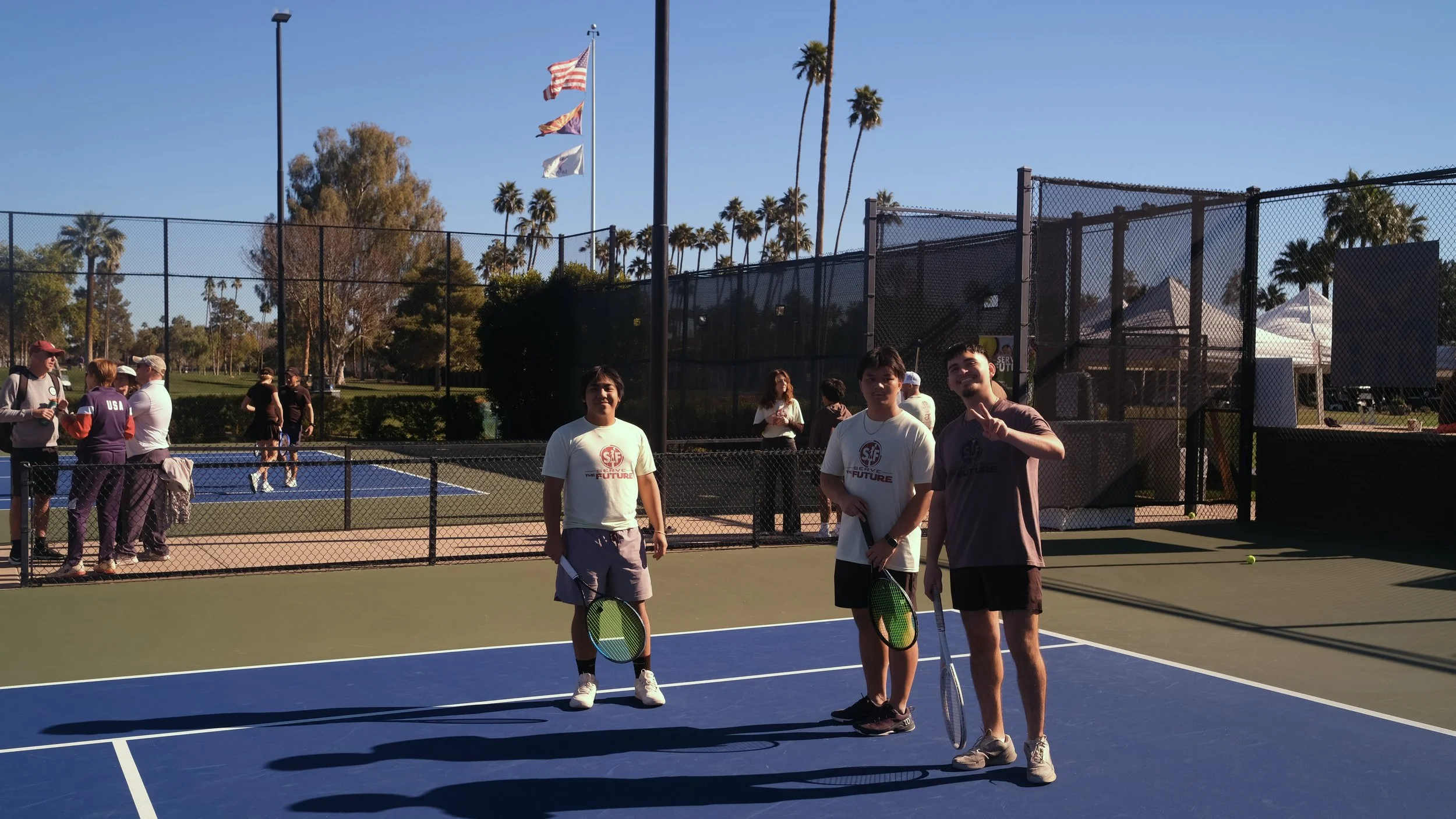 People standing on a tennis court holding paddles, with children and adults in the background, surrounded by palm trees and flags.