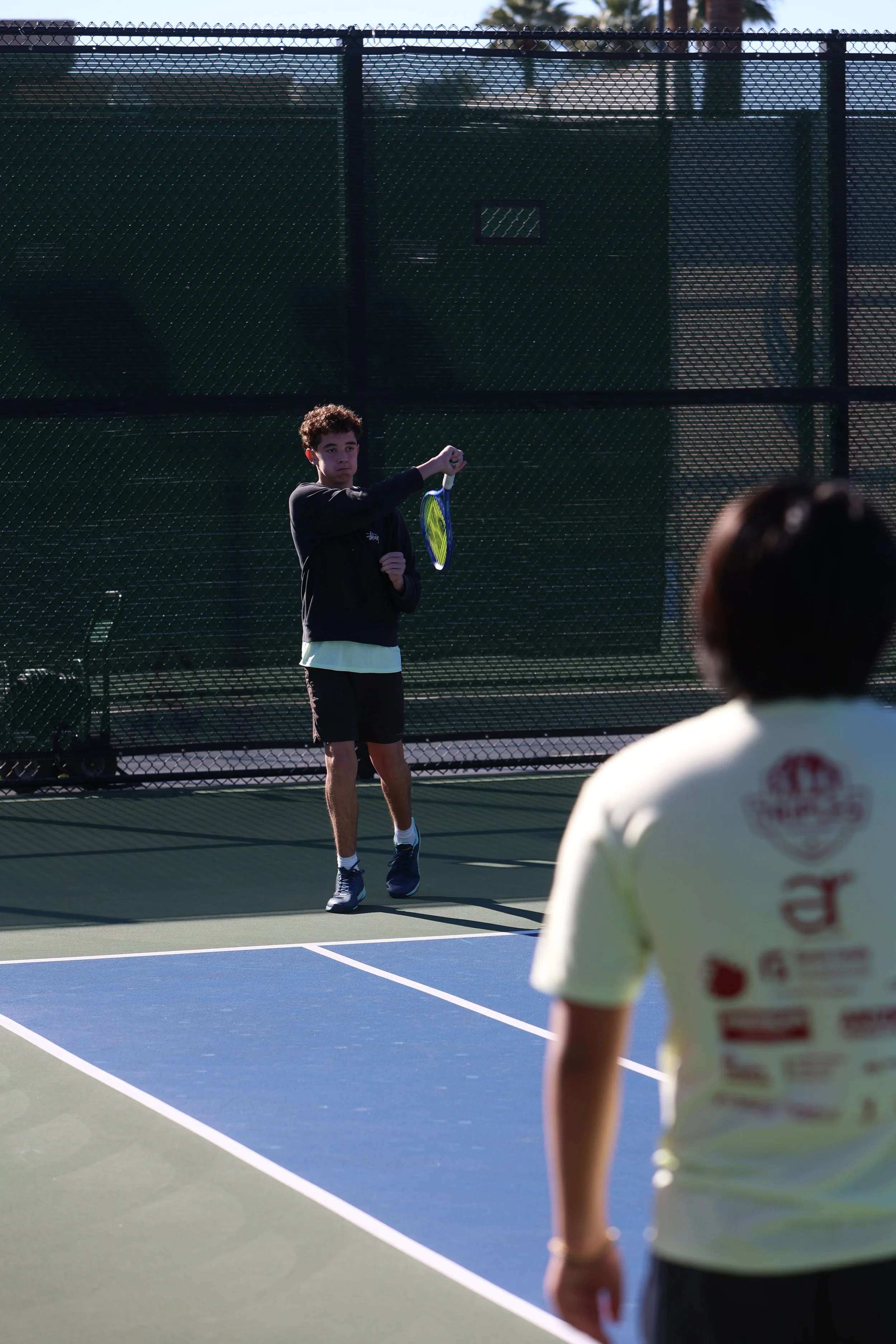 Two boys playing tennis on an outdoor court, with one boy preparing to hit the ball and the other watching.