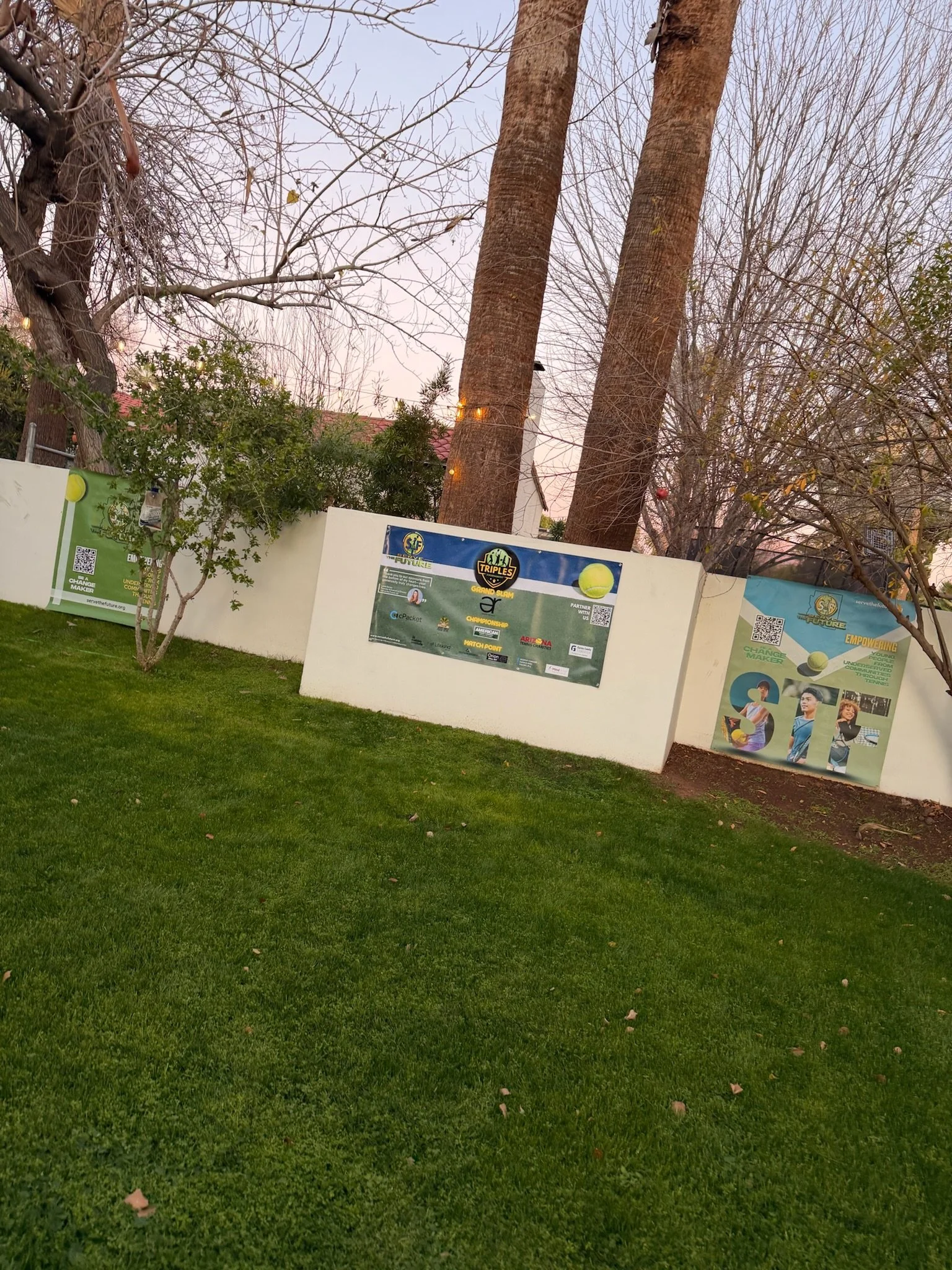 A white wall with posters and banners on it, surrounded by trees with bare branches and small green trees, with bright green grass in the foreground.
