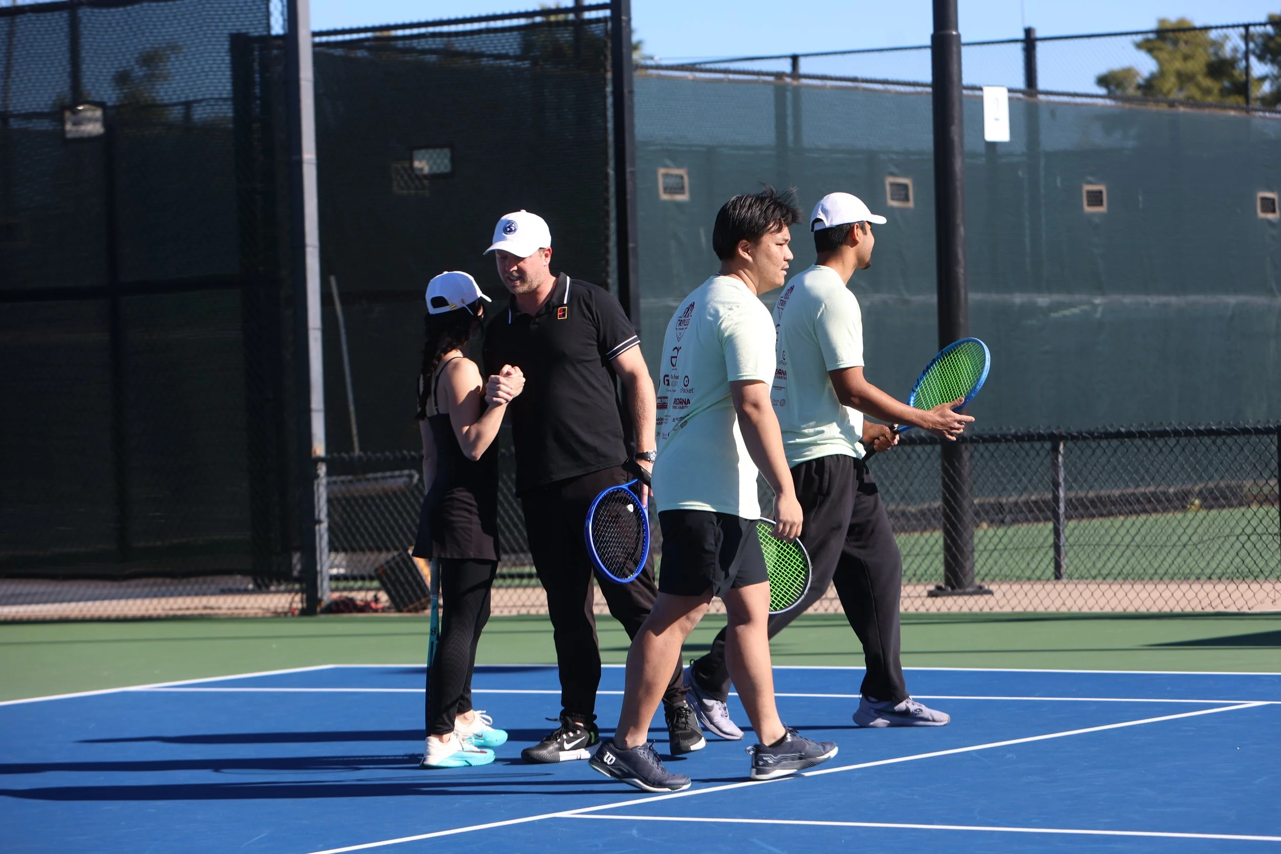 Four people on a tennis court, coaching or instructing each other, with two of them holding tennis rackets and wearing tennis apparel, the background showing a green fence and a clear sky.