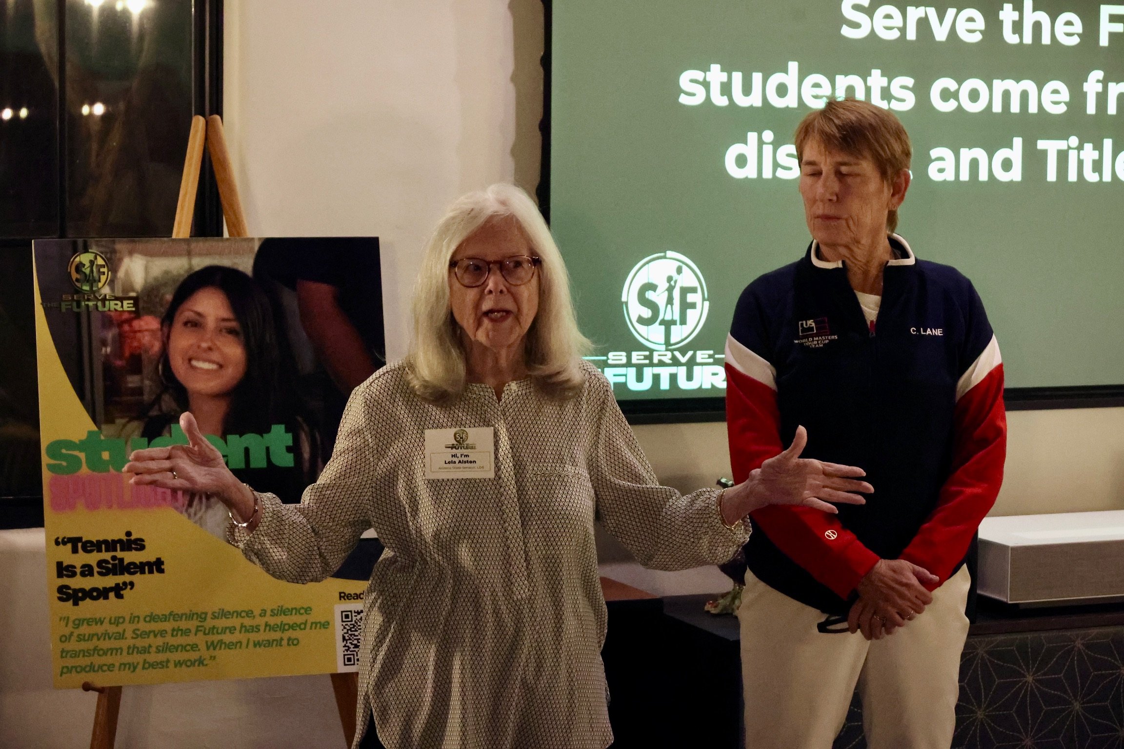 Three women standing indoors, one speaking and gesturing with her hands. Behind them is a large screen with text about serving students and a logo that says 'Serve the Future.' To the left, there is a poster with a photo of a woman, the phrase 'Tenni