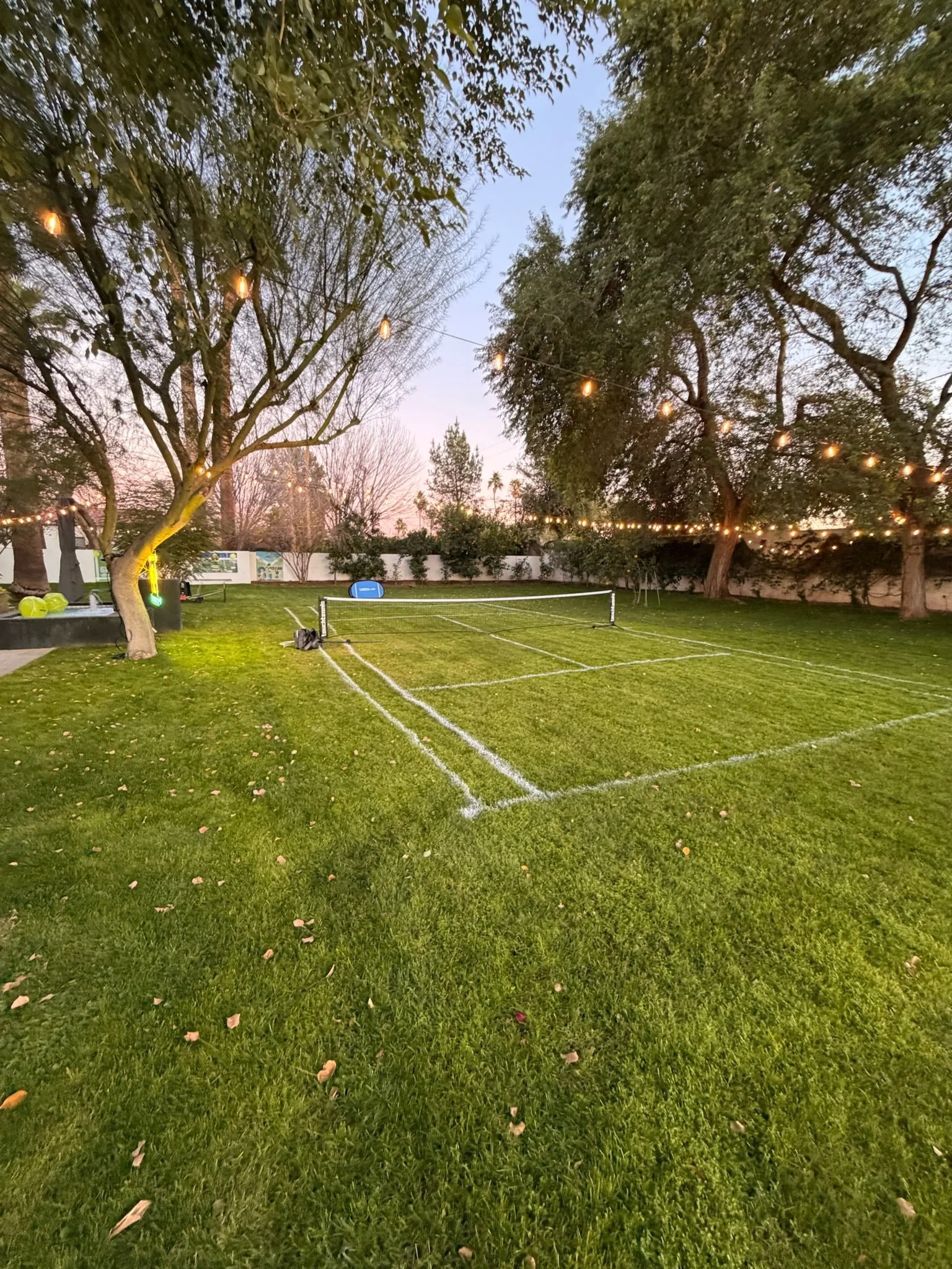 An outdoor backyard with tennis courts, surrounded by trees with string lights hanging overhead at sunset or dusk.