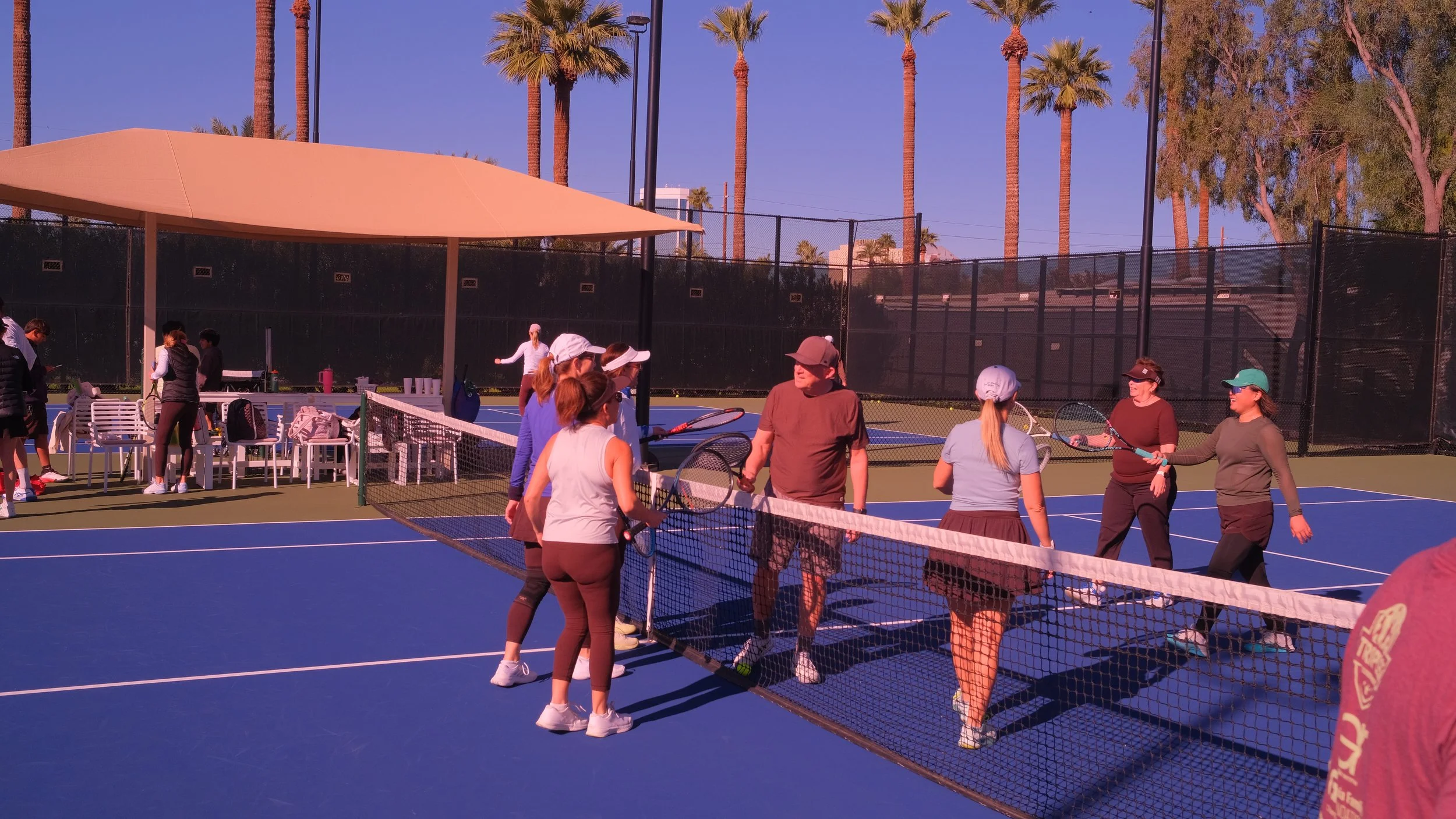 People on a tennis court participating in a tennis lesson or group activity, with a net, blue surface, and palm trees in the background under a blue sky.