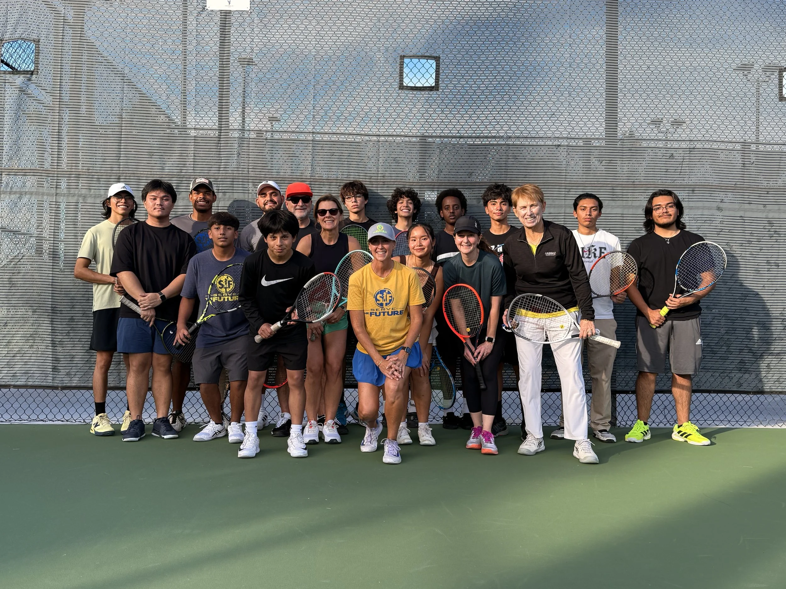 Group of tennis players and coaches posing on a tennis court after a match, holding tennis rackets.