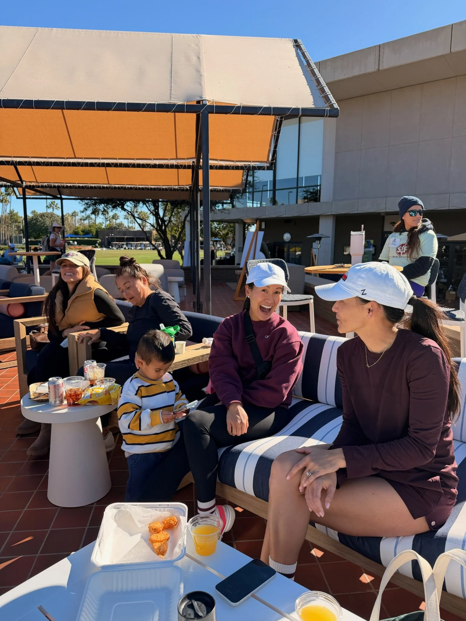 Group of women and children sitting and chatting outdoors on a patio with tables, drinks, and snacks, under a large orange canopy, with modern buildings and trees in the background on a sunny day.