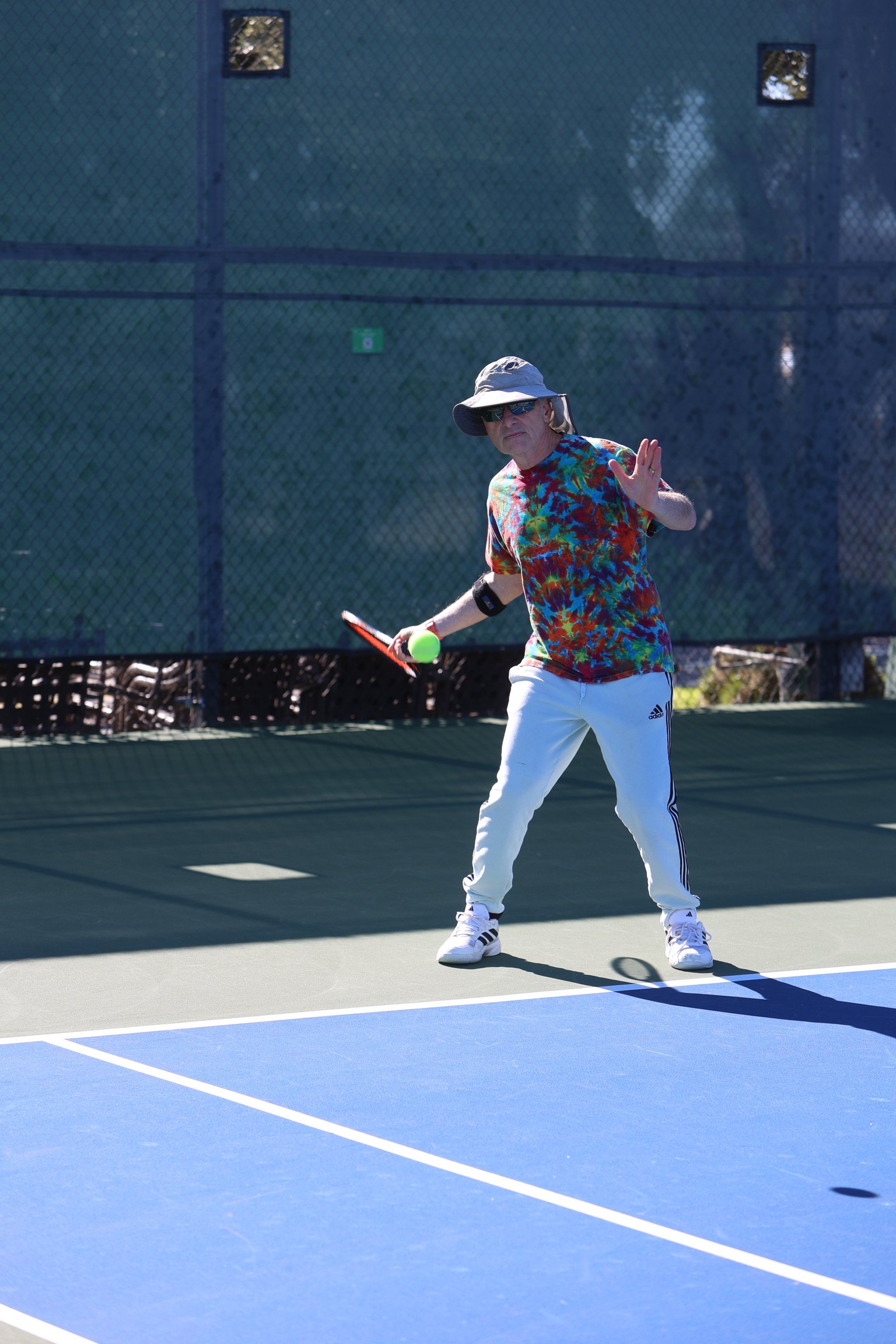 A person playing tennis on an outdoor court, wearing a colorful tie-dye shirt, white pants, a wide-brimmed hat, sunglasses, and holding a tennis racket while preparing to hit a tennis ball.