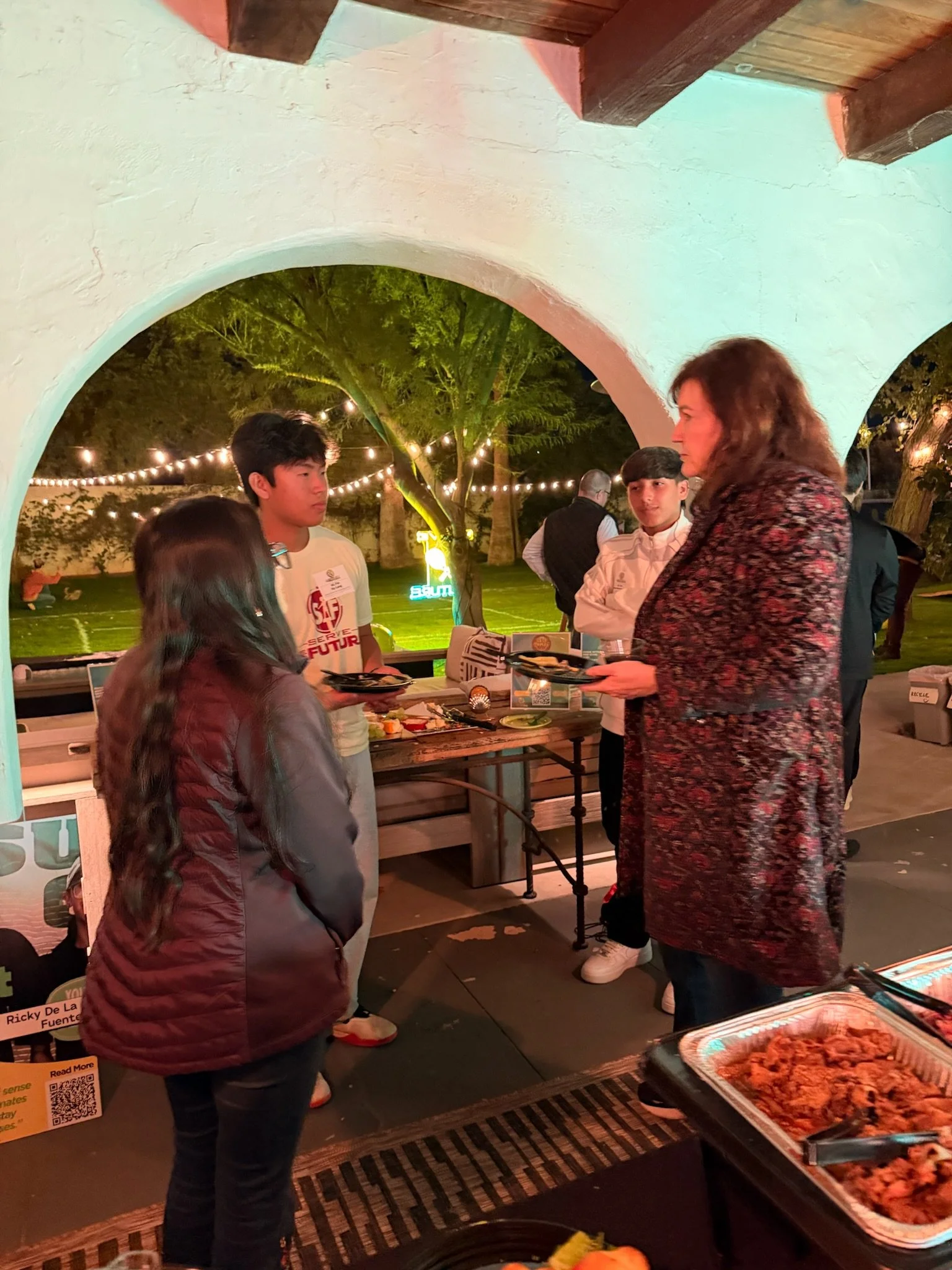People enjoying an outdoor evening gathering with food, under hanging string lights and trees.