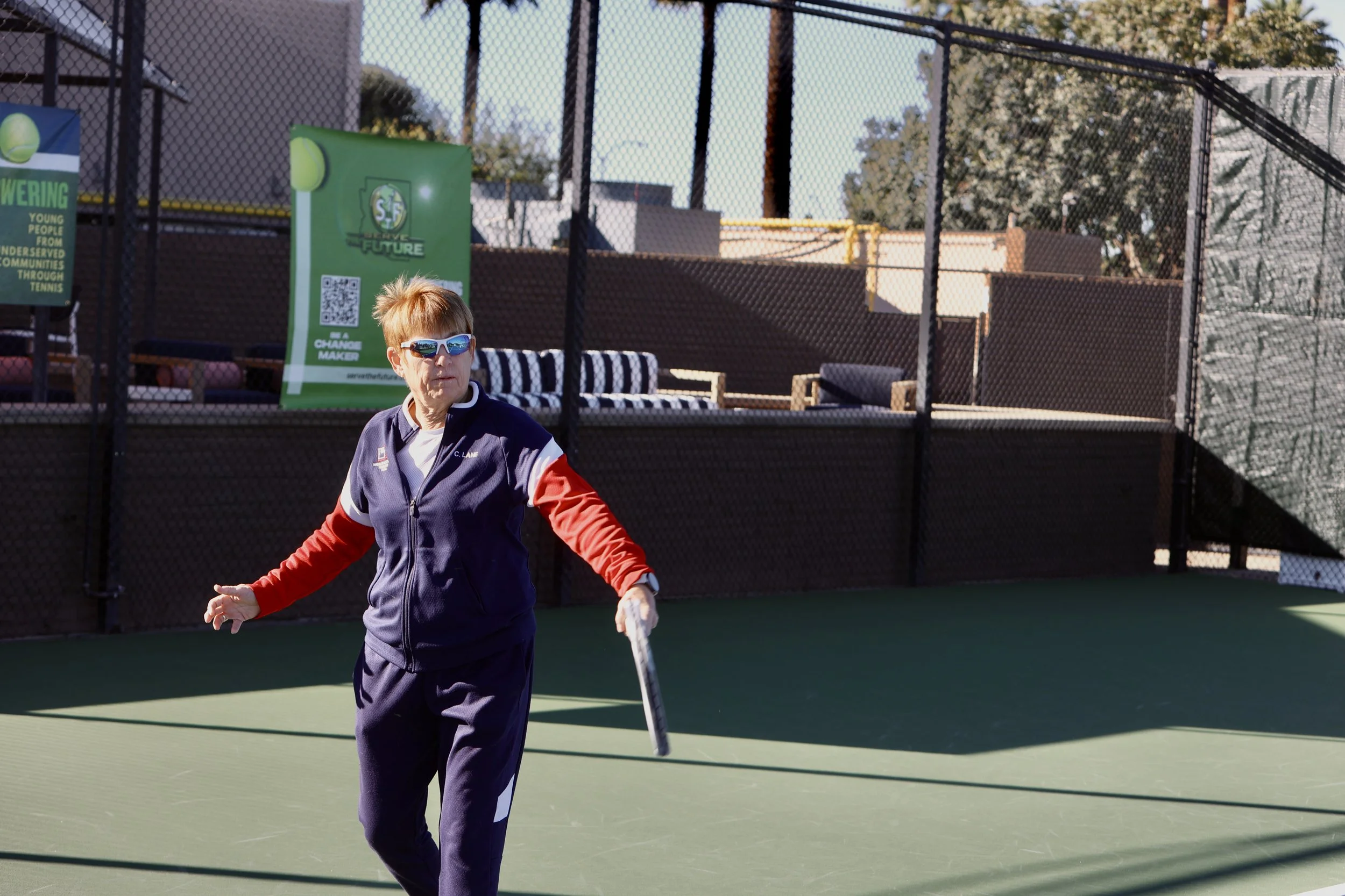 An elderly woman with short hair wearing sunglasses and athletic clothing, holding a pickleball paddle on a pickleball court with a chain-link fence and outdoor seating in the background.