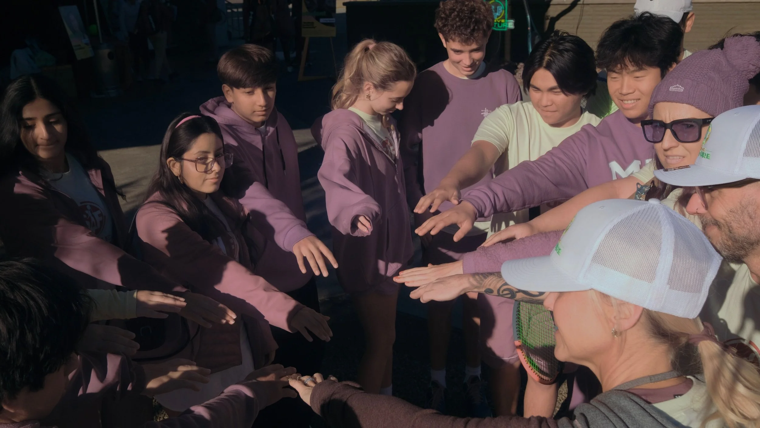 A group of teenagers and adults with diverse ethnicities gathering outdoors, participating in a team-building activity where they extend their hands together in a circle, engaging in a shared moment of camaraderie.