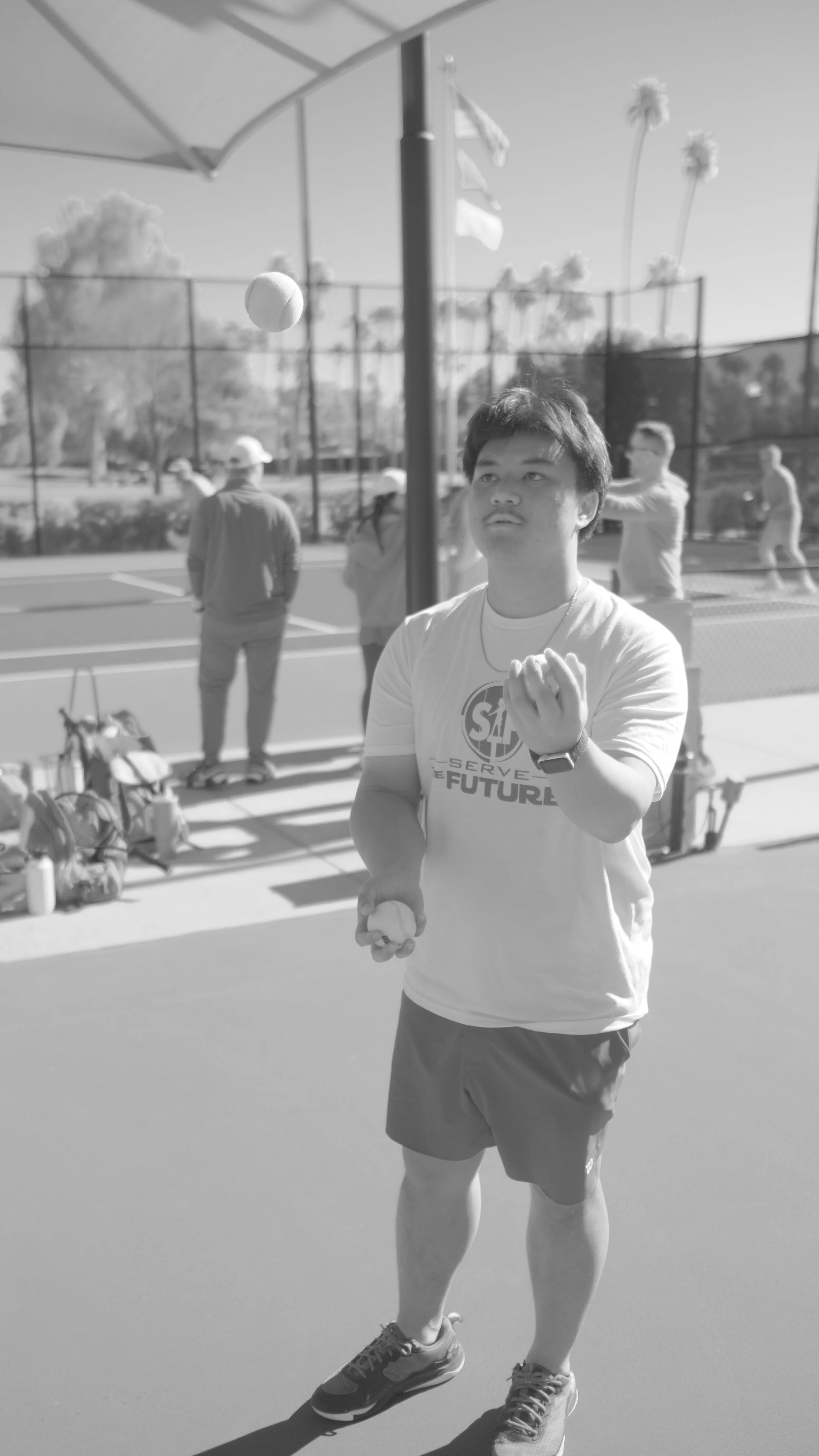 A young man with dark hair, wearing a t-shirt that says 'Serve the Future,' shorts, and sneakers, is holding a tennis ball in one hand and preparing to serve on a tennis court. In the background, several people are standing or preparing to play, with
