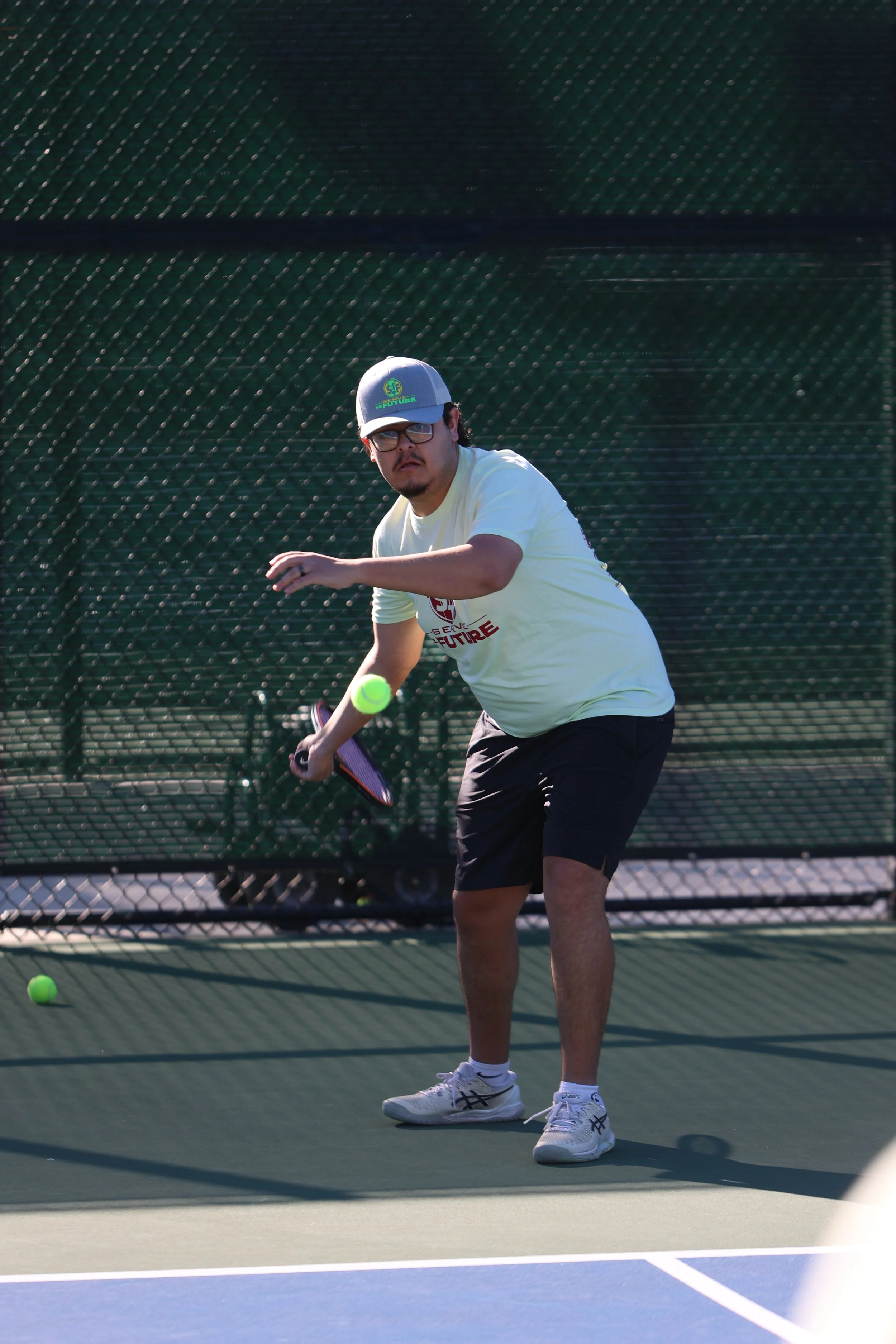 Man wearing glasses, a cap, and a light yellow T-shirt playing tennis on an outdoor court, preparing to hit a tennis ball with a racket.