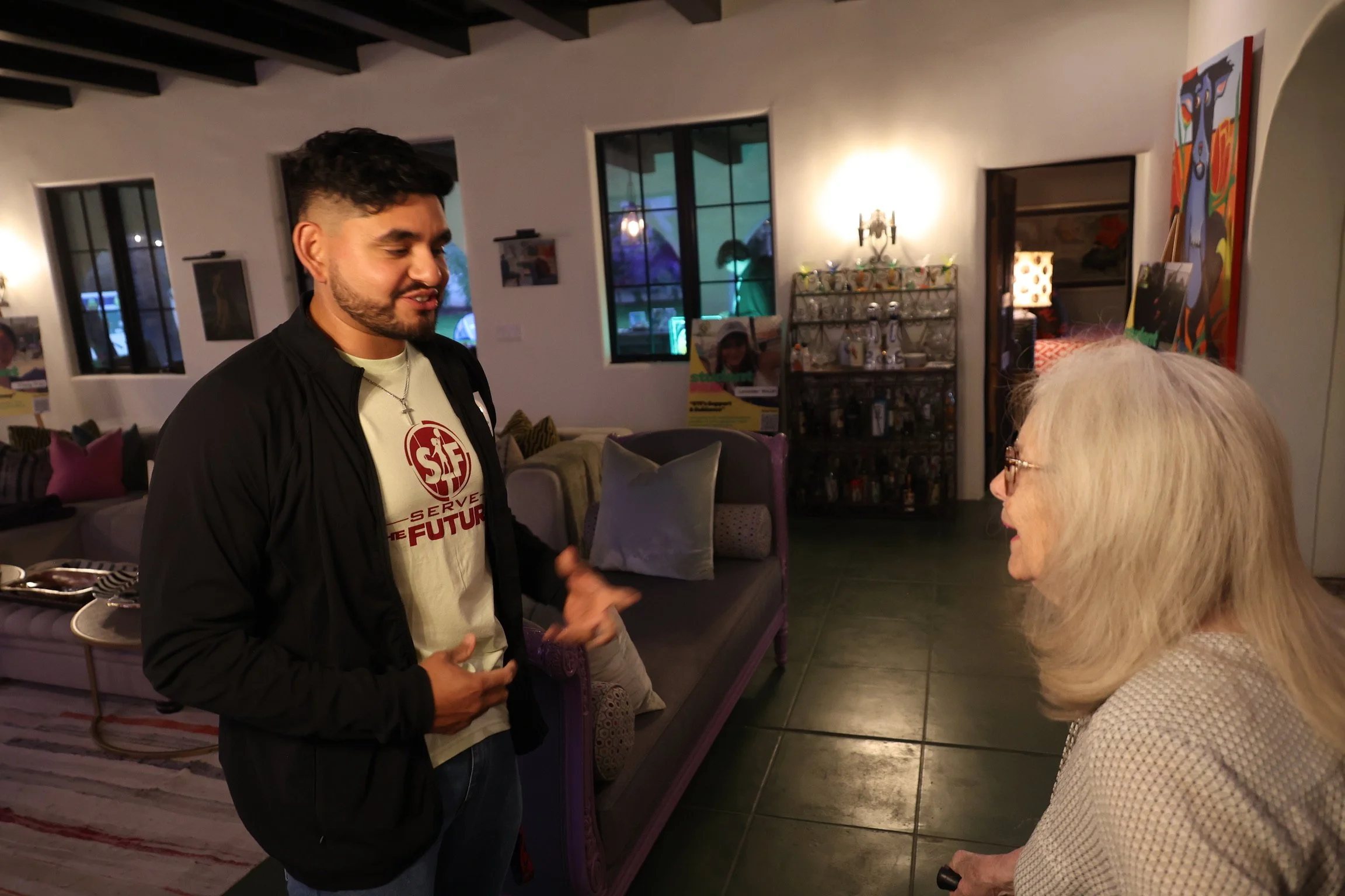 A young man with dark hair and a beard talking to an elderly woman with long white hair in a cozy, decorated living room with artwork on the walls, a bar cart, and windows with a nighttime view.