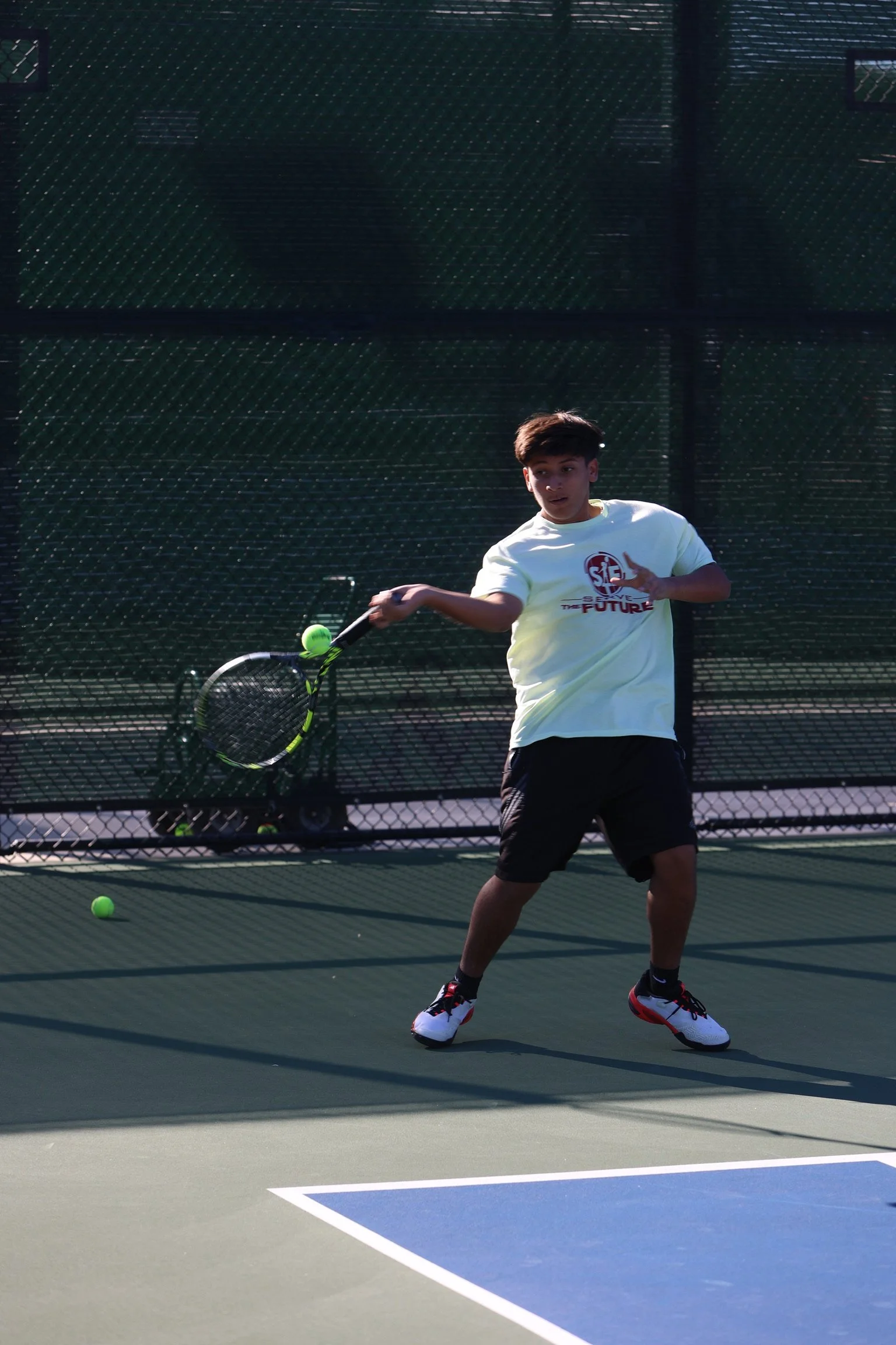 A young male tennis player in a white T-shirt and black shorts hitting a tennis ball with a racket on an outdoor tennis court, with a fence in the background.