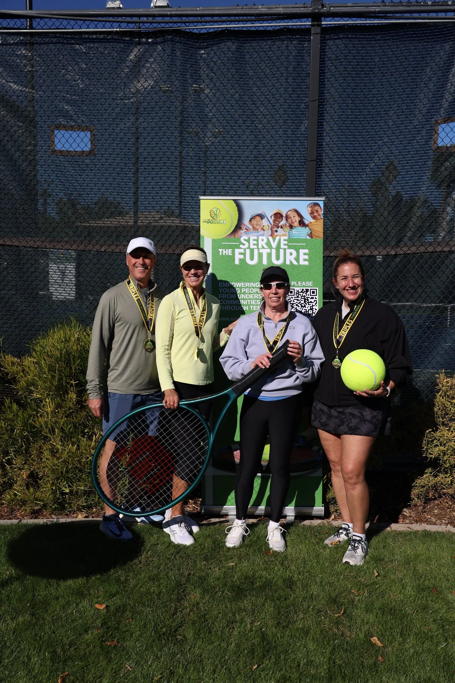 Four women standing outdoors in front of a green and white checkered fence, holding tennis equipment and a tennis ball, with a tennis-themed banner that reads "Serve the Future" behind them.