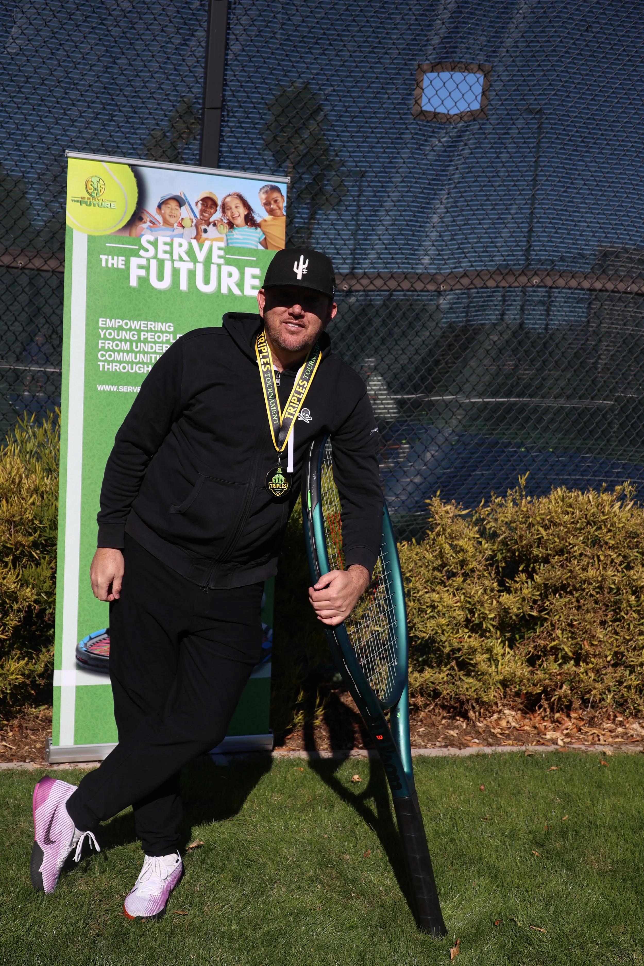 Man holding tennis racket standing in front of a "Serve the Future" sign on a tennis court.