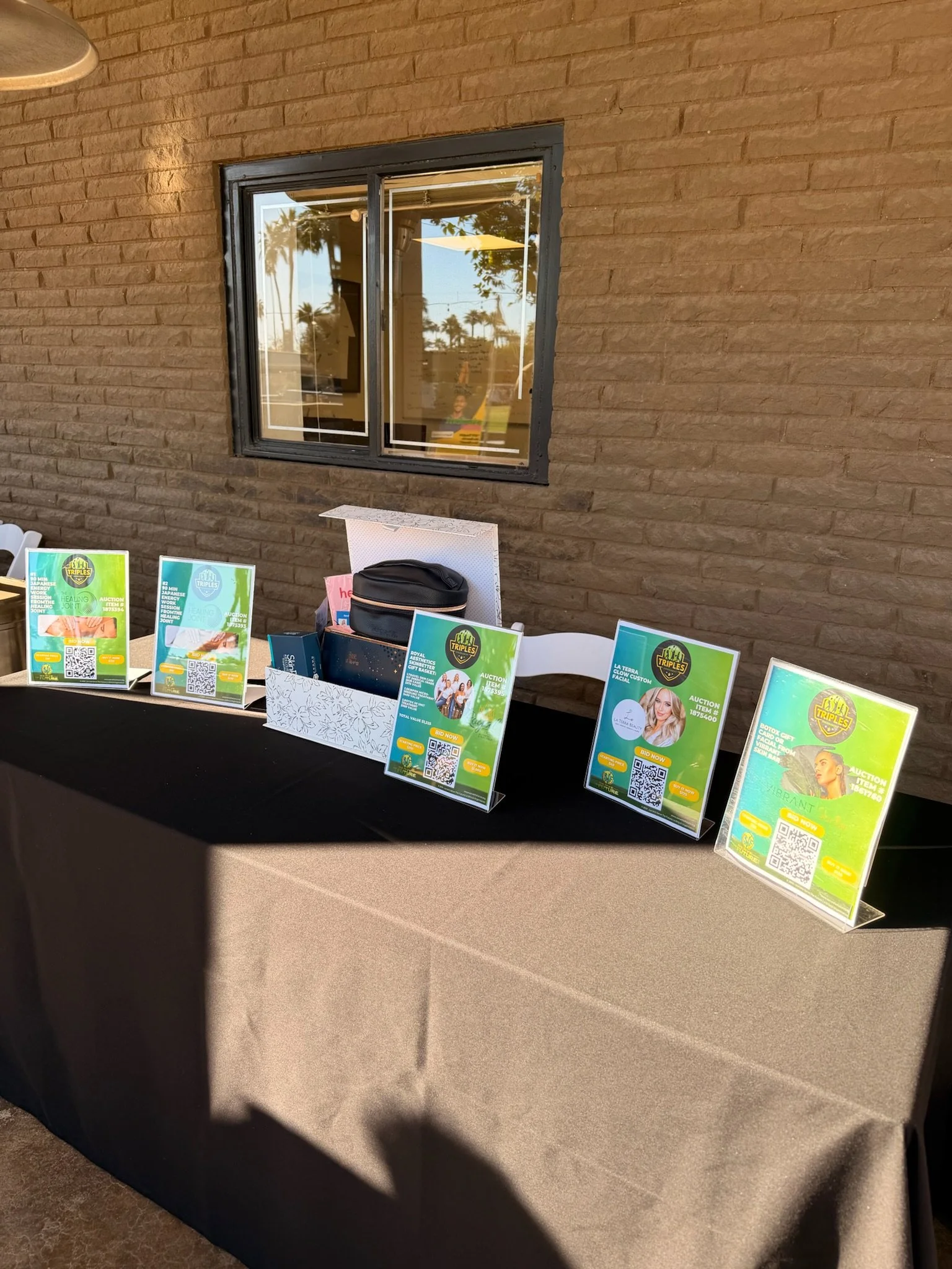 A black tablecloth-covered table with posters for an auction, a black bag, and a white gift box, set against a brick wall with a window reflecting outdoor trees and sky.