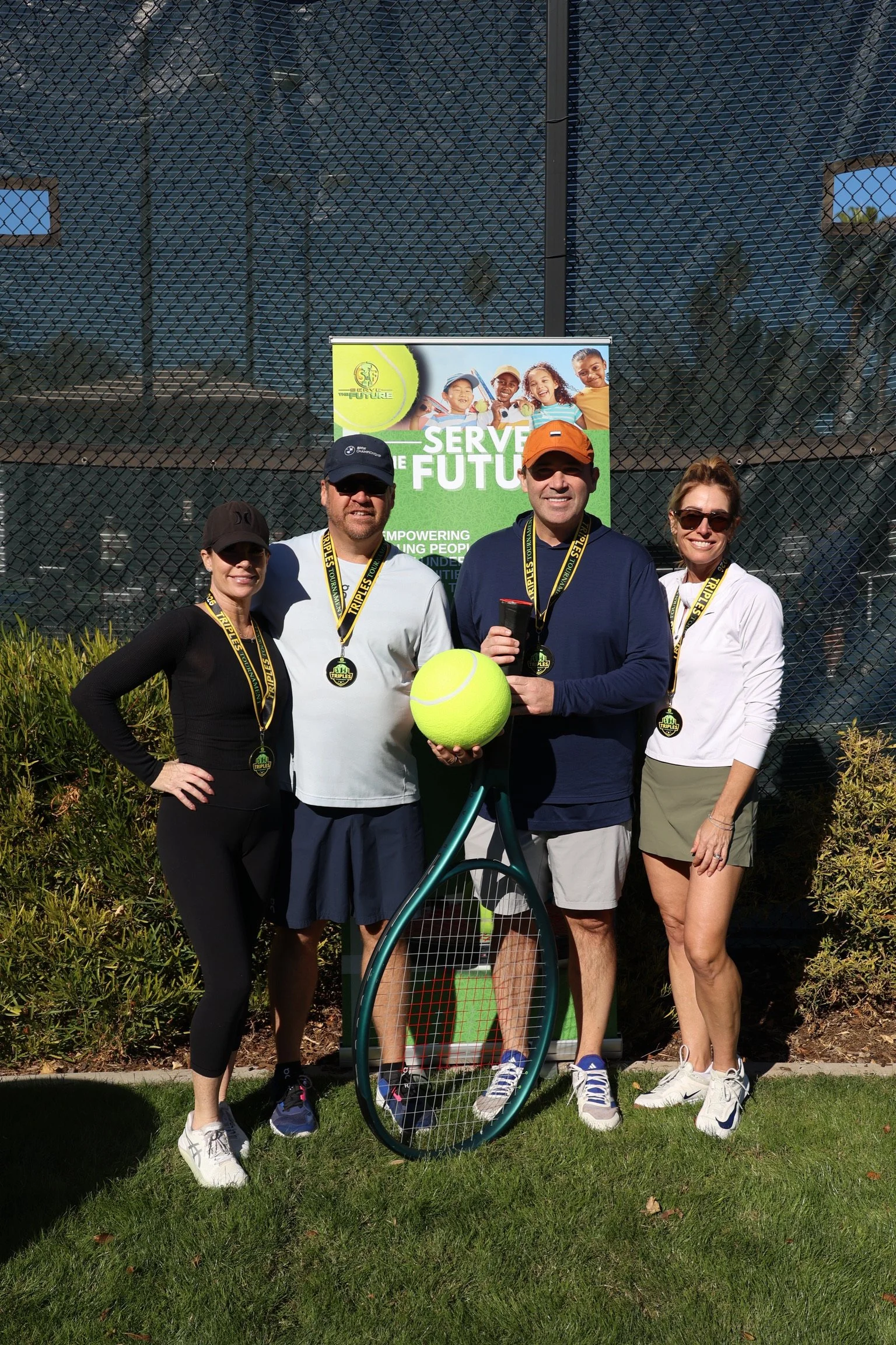 Four people standing outdoors in front of a sign that reads 'Serve the Future'. They are wearing medals around their necks, and one person is holding a tennis ball and a tennis racket. The group is smiling, and there is greenery and a tennis court fe