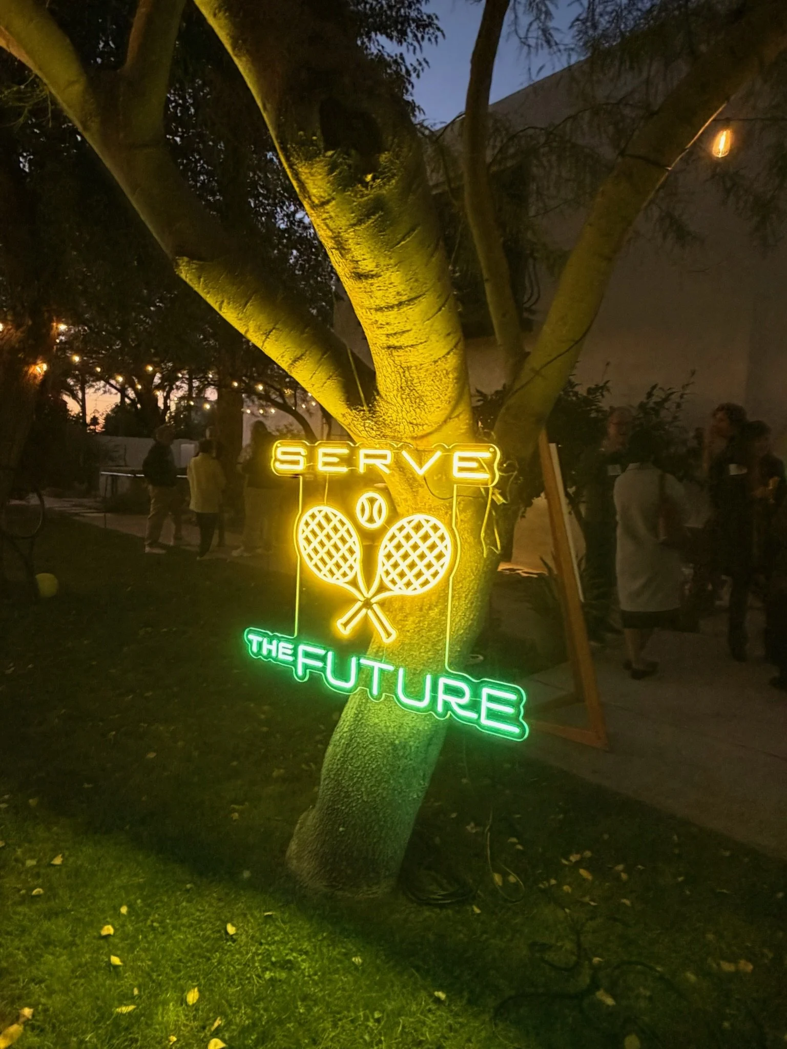Neon sign hanging from a tree at dusk that reads 'Serve the Future' with two crossed tennis rackets and a tennis ball.