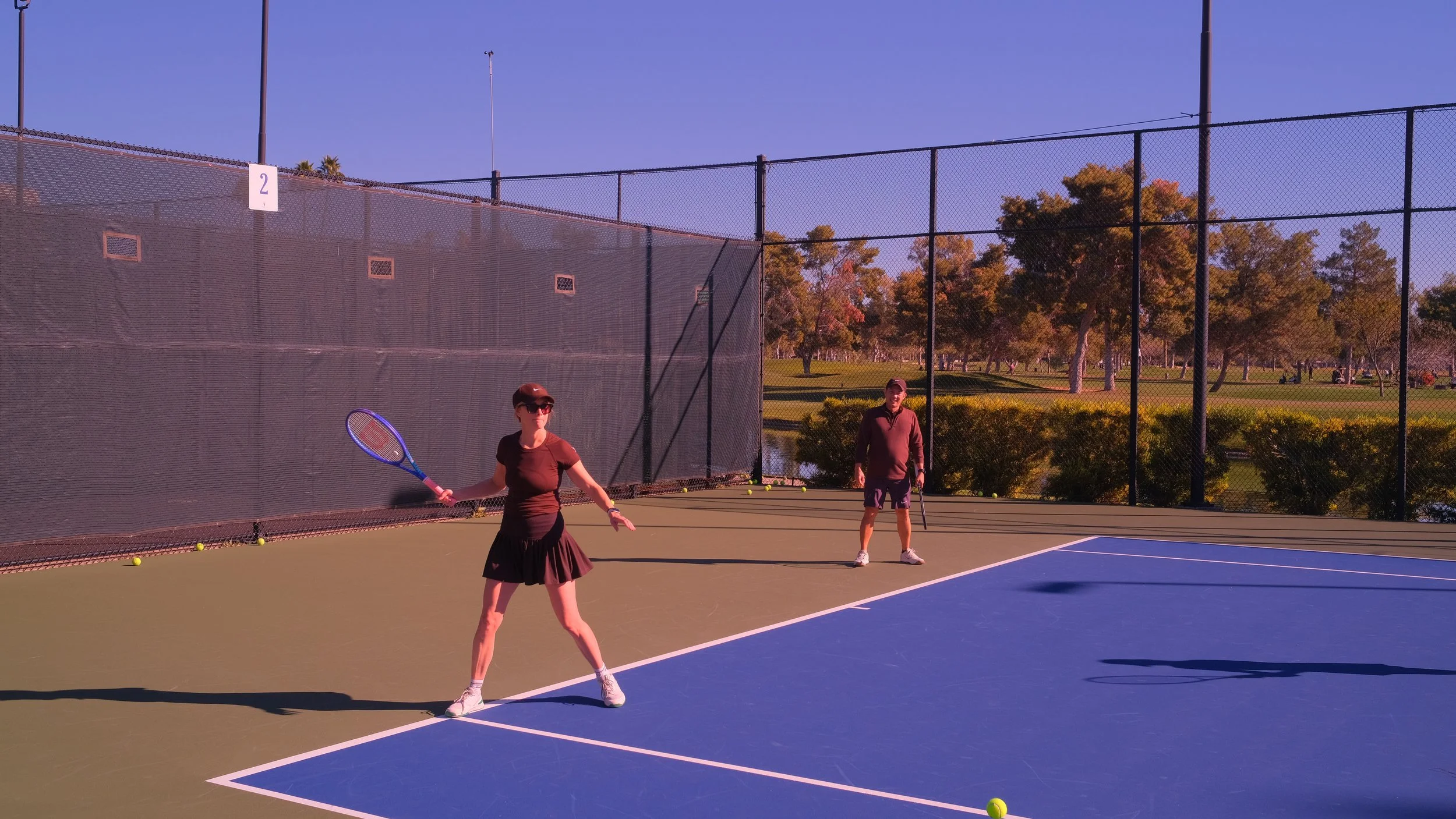 Two people practicing tennis on an outdoor court during daytime. The woman in the foreground is wearing a black skirt, shirt, visor, and sunglasses, holding a tennis racket. The man in the background is dressed in a dark jacket and shorts, standing n