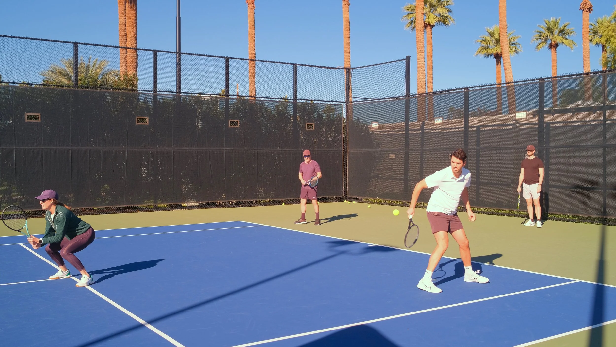 Four people playing tennis on an outdoor court with tall palm trees in the background.