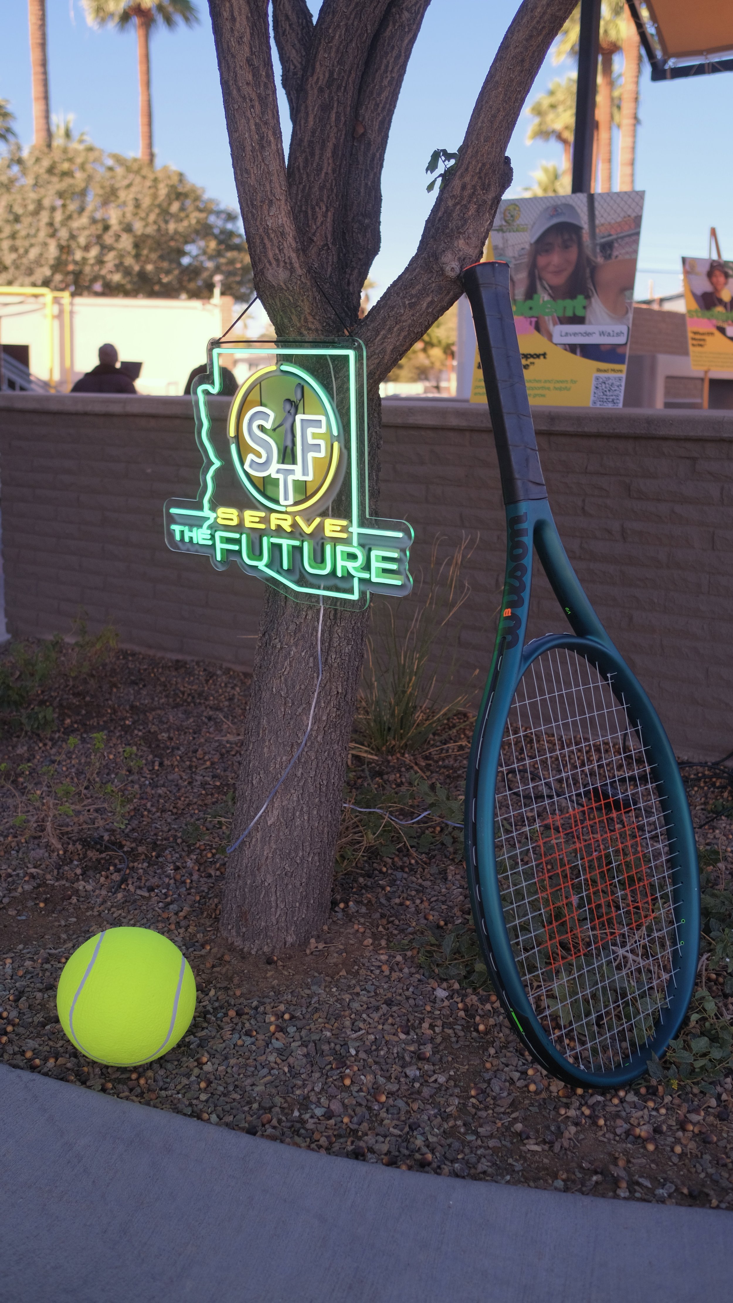 A neon sign hanging on a tree reads, 'SF Serve the Future.' A yellow tennis ball is on the ground nearby, and a tennis racket leans against the tree. In the background, there are posters and a brick wall.