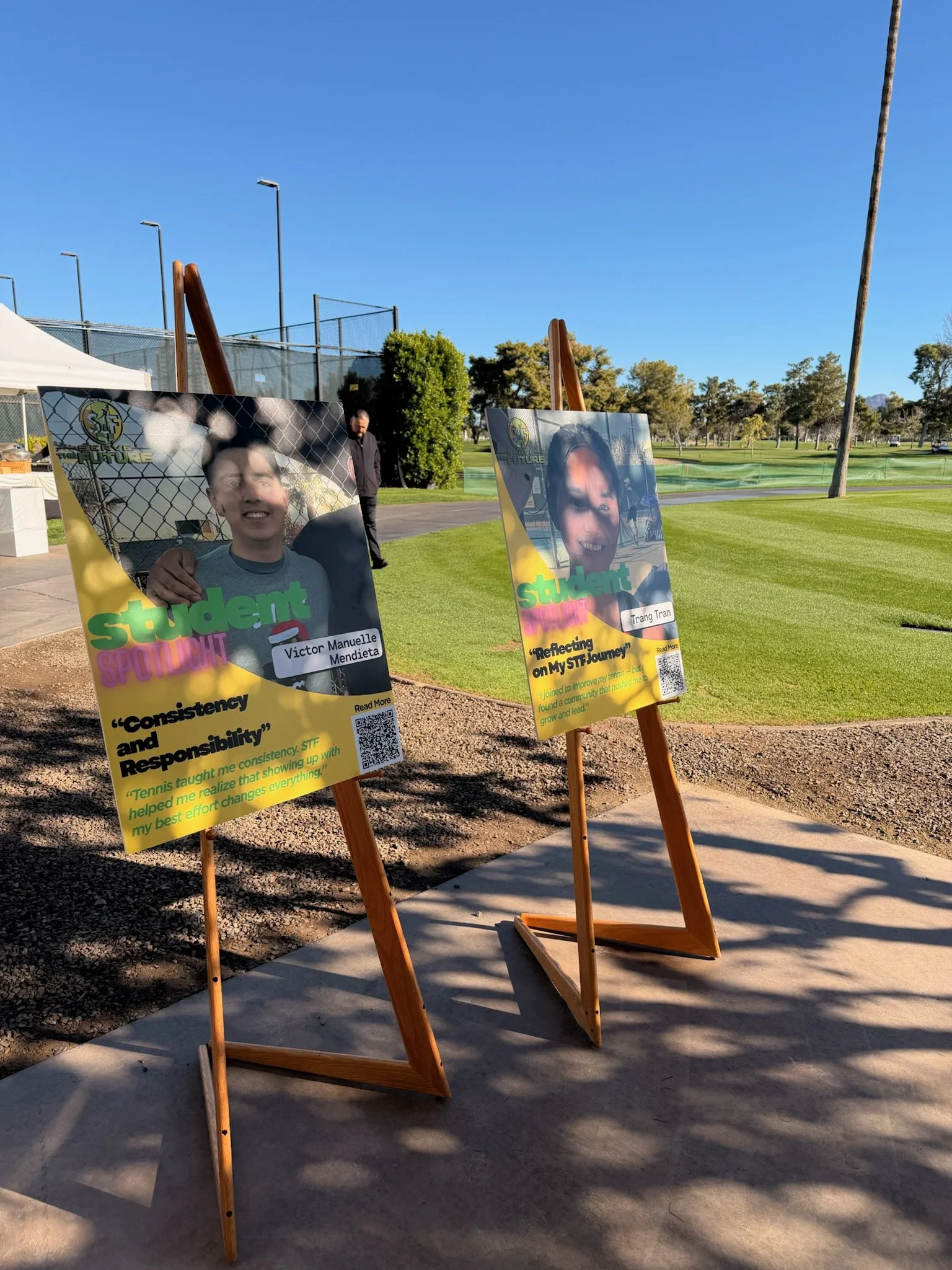Two colorful outdooor posters on easels featuring students' photos and quotes, set up near a grassy area on a sunny day, with tennis courts, trees, and blue sky in the background.