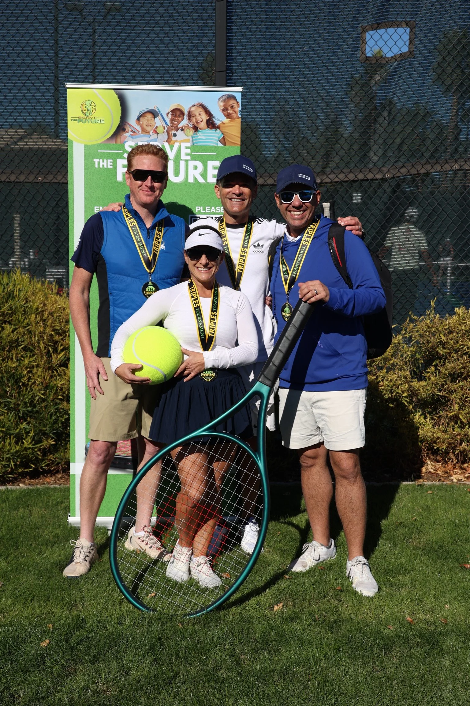 Four people standing outdoors in front of a tennis court, smiling and wearing medals, holding a tennis racket and a large tennis ball. They are dressed in sportswear, and there is a green poster behind them that reads "Save the Future" with a group o
