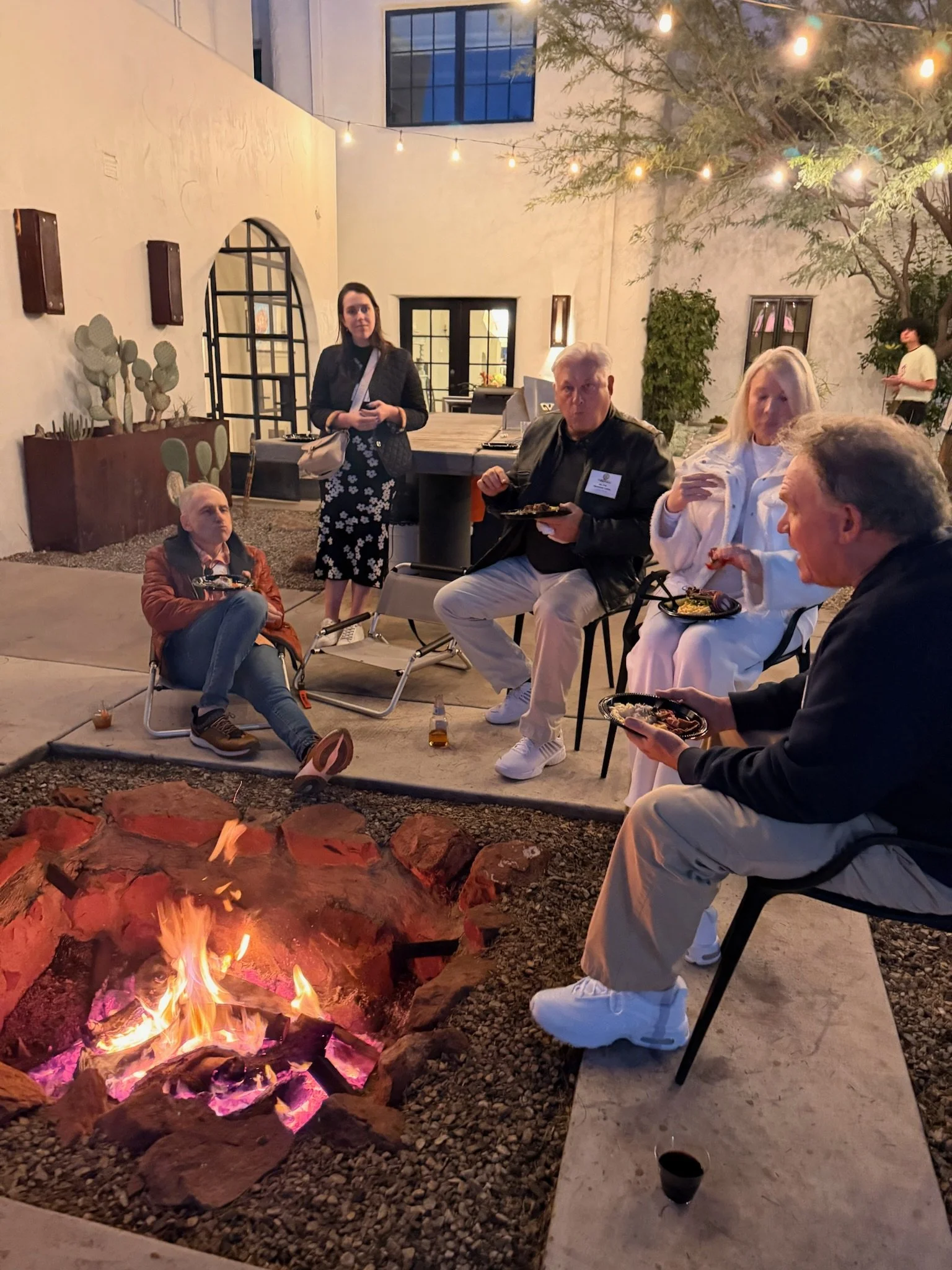 People sitting around a fire pit in an outdoor courtyard, enjoying food and drinks at night. String lights are hanging overhead, and a few people are standing in the background.