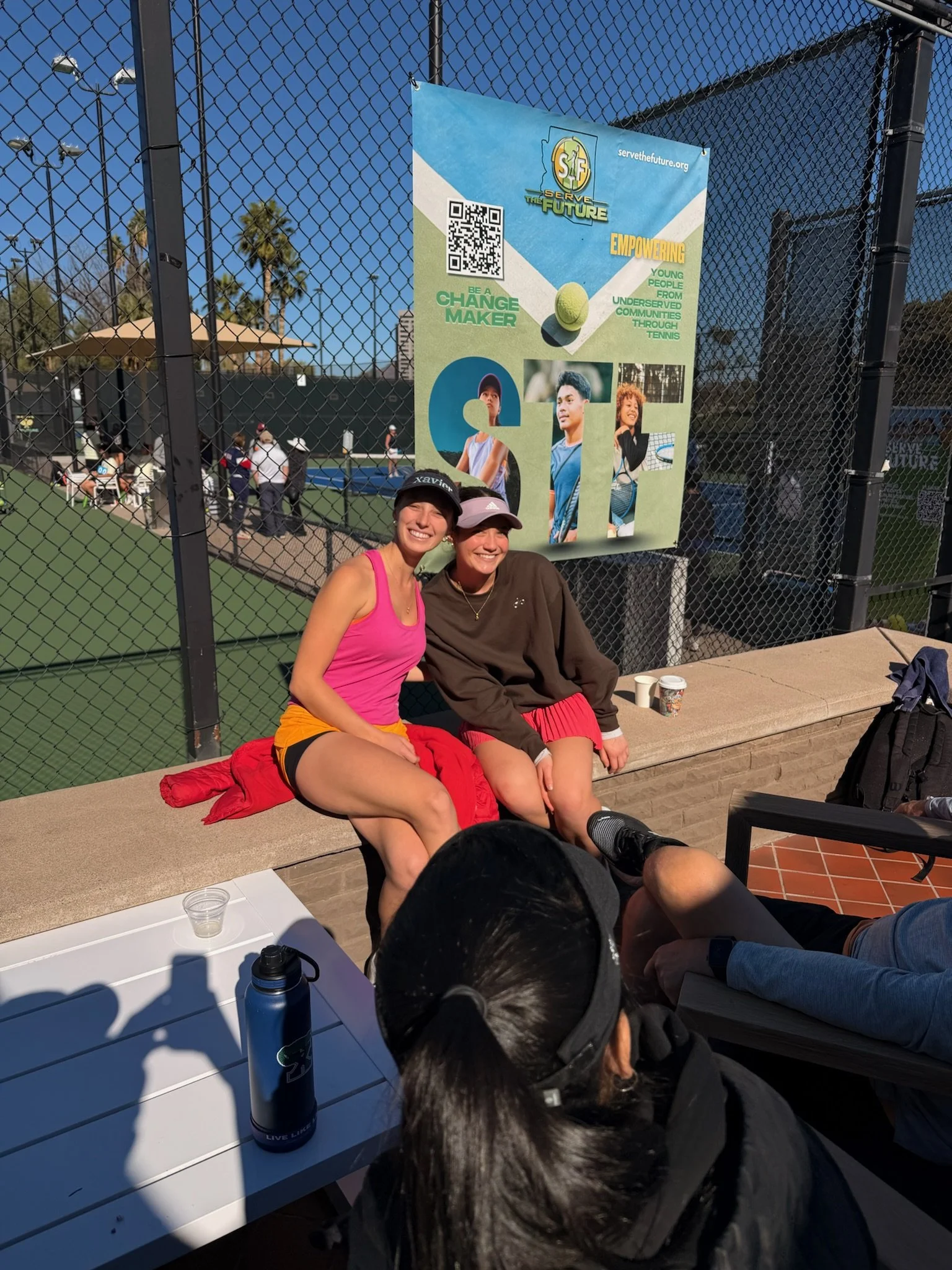 Two smiling women sitting on a bench at an outdoor tennis court; one wearing a pink tank top and yellow shorts, the other in a brown sweatshirt and red shorts, with people playing tennis in the background and a large promotional banner for Serve the 