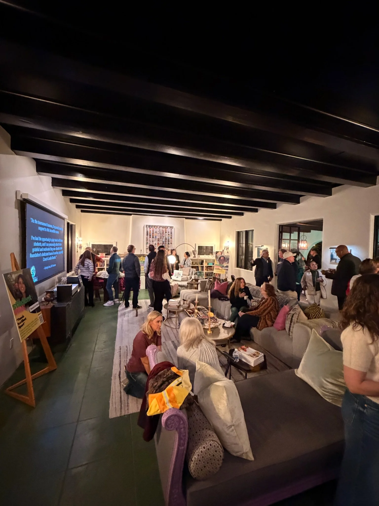 Indoor gathering with people seated on sofas and standing, in a warmly lit room with black ceiling beams, large windows, and various artworks and bookshelves on the walls.