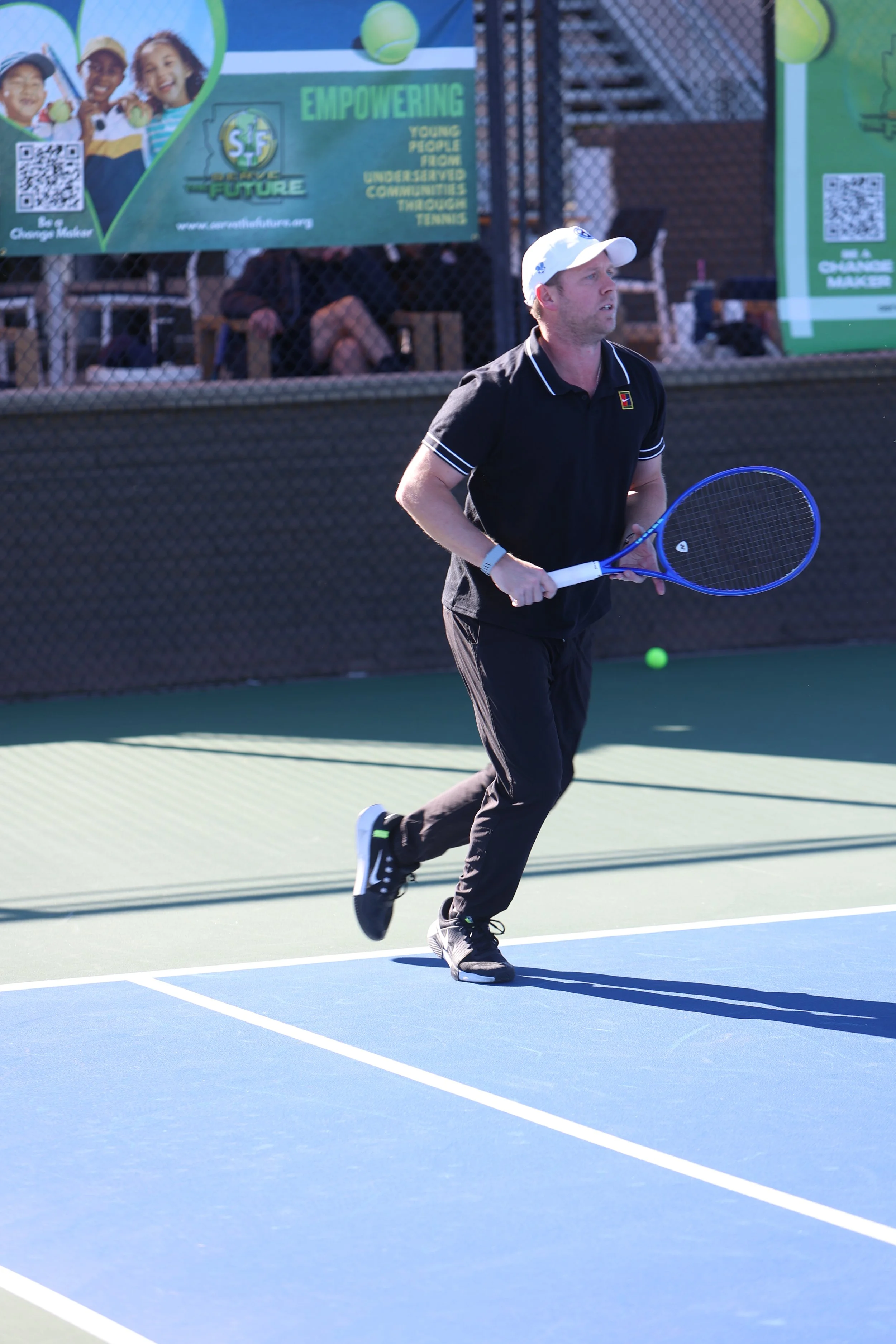A man playing tennis on a court, holding a tennis racket, with a ball nearby, during daytime, in front of a fence with advertisements.