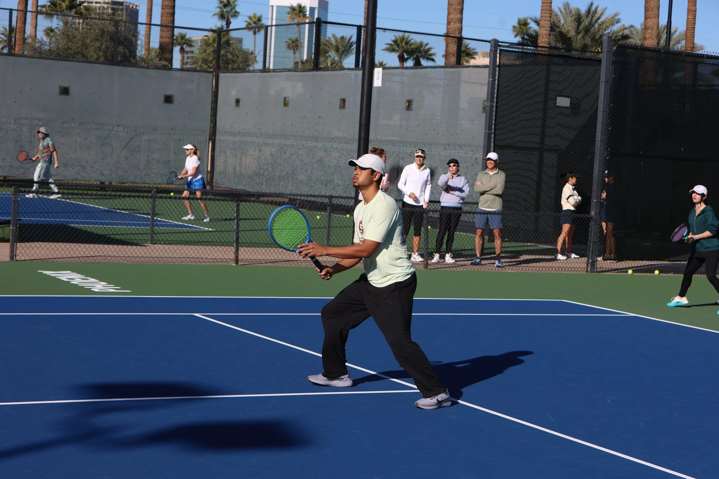 Young man playing tennis on a blue court with a green area in the background. Several people watch from behind a fence and some are also playing on nearby courts. Palm trees and a clear sky are visible in the background.