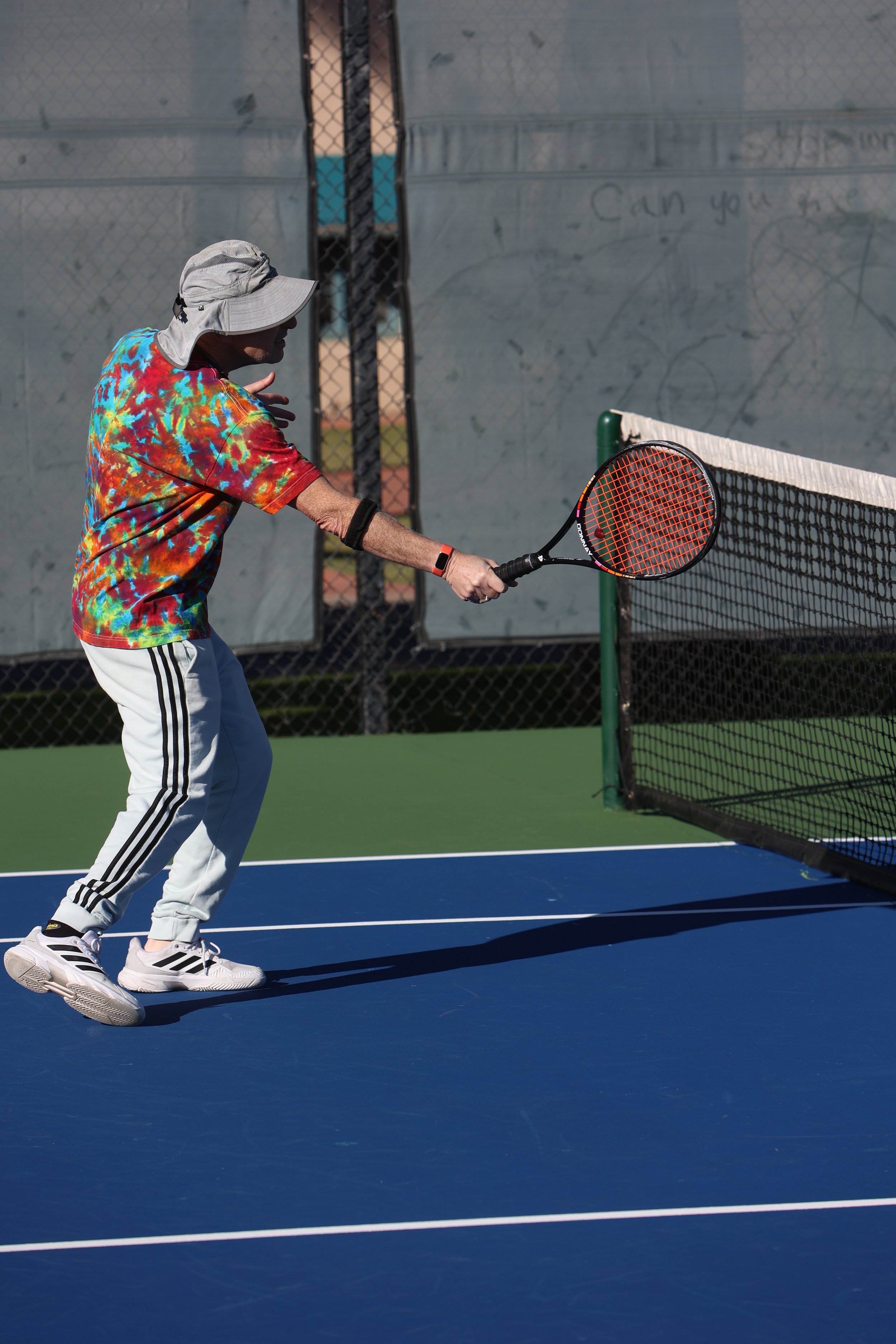 A man wearing a tie-dye shirt, white pants with black stripes, and a hat playing tennis on a court, preparing to hit a ball with a tennis racket.