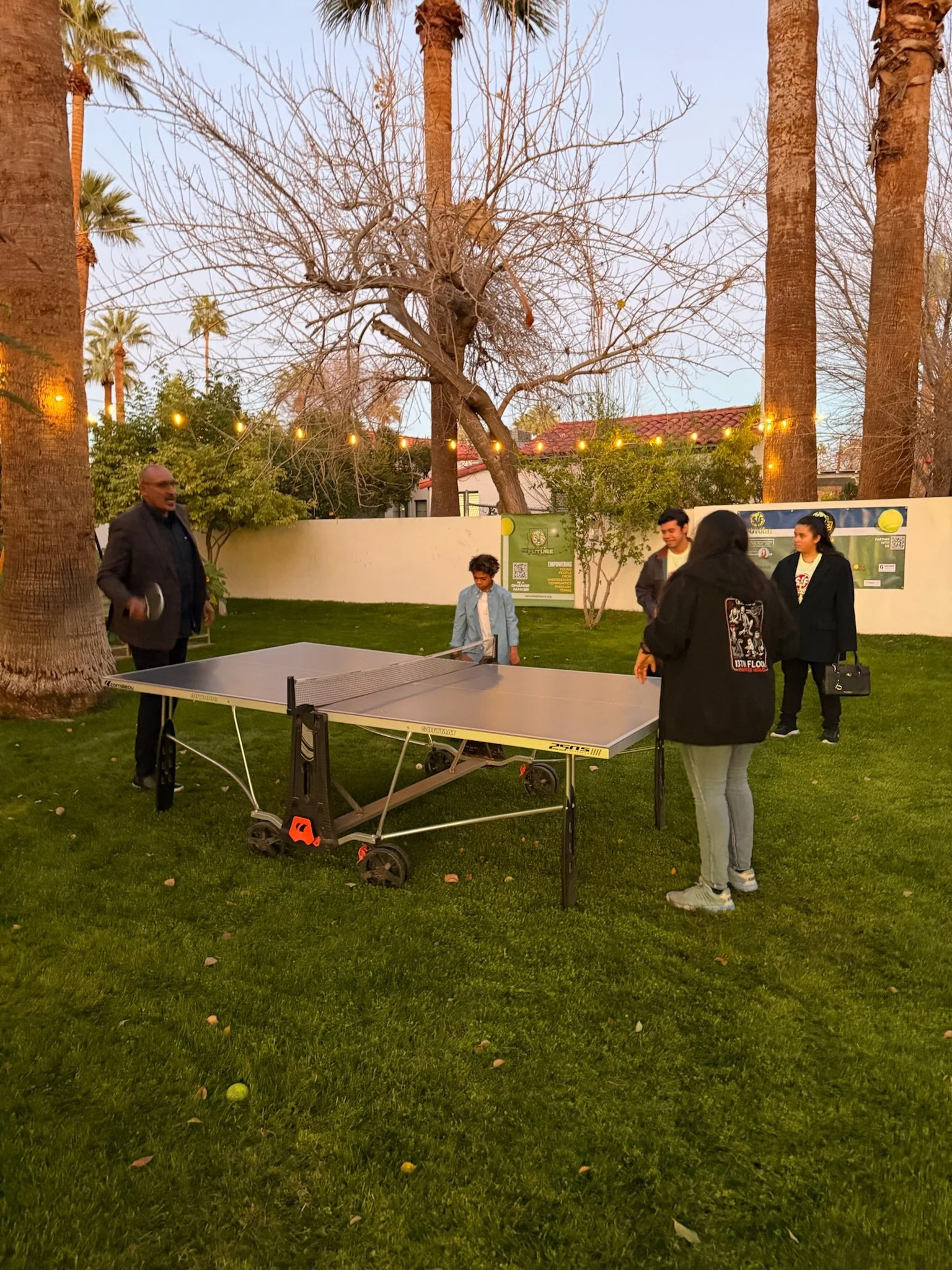 People playing ping pong outdoors in a backyard with large trees, string lights, and a green lawn at dusk.