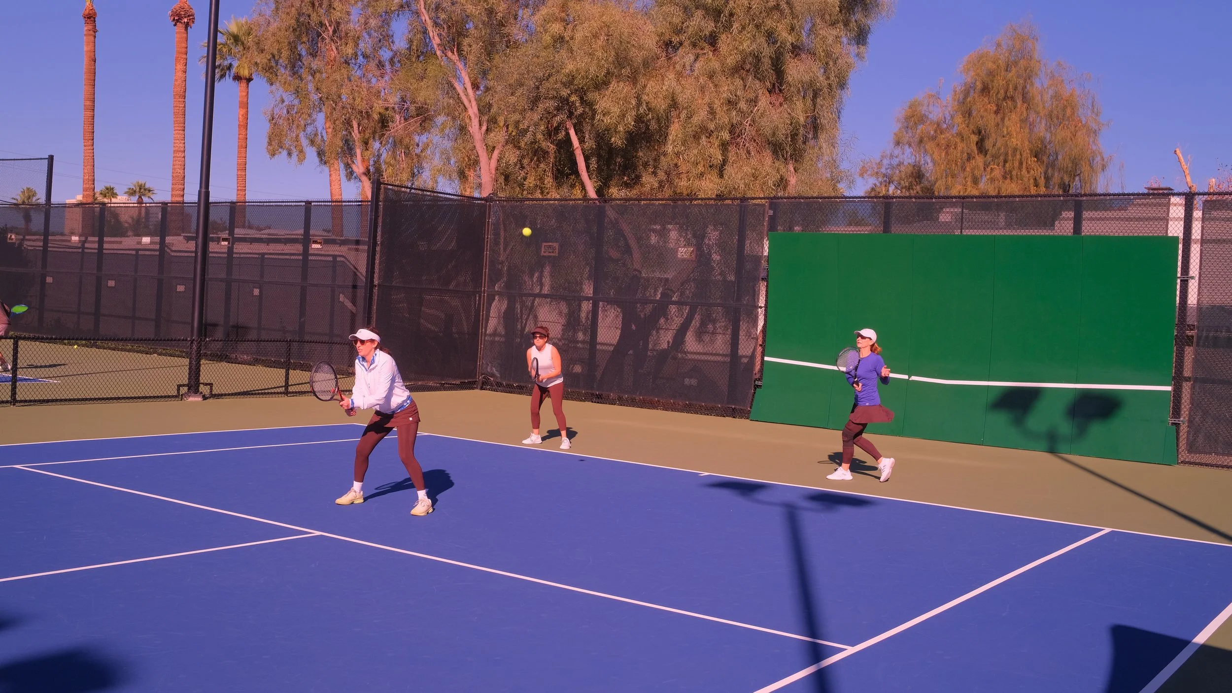 Three women playing pickleball on a blue and green outdoor court. Two women are positioned near the net, preparing for a shot, while the third woman is standing further back. The court is enclosed by a black fence, and there are tall palm trees and o