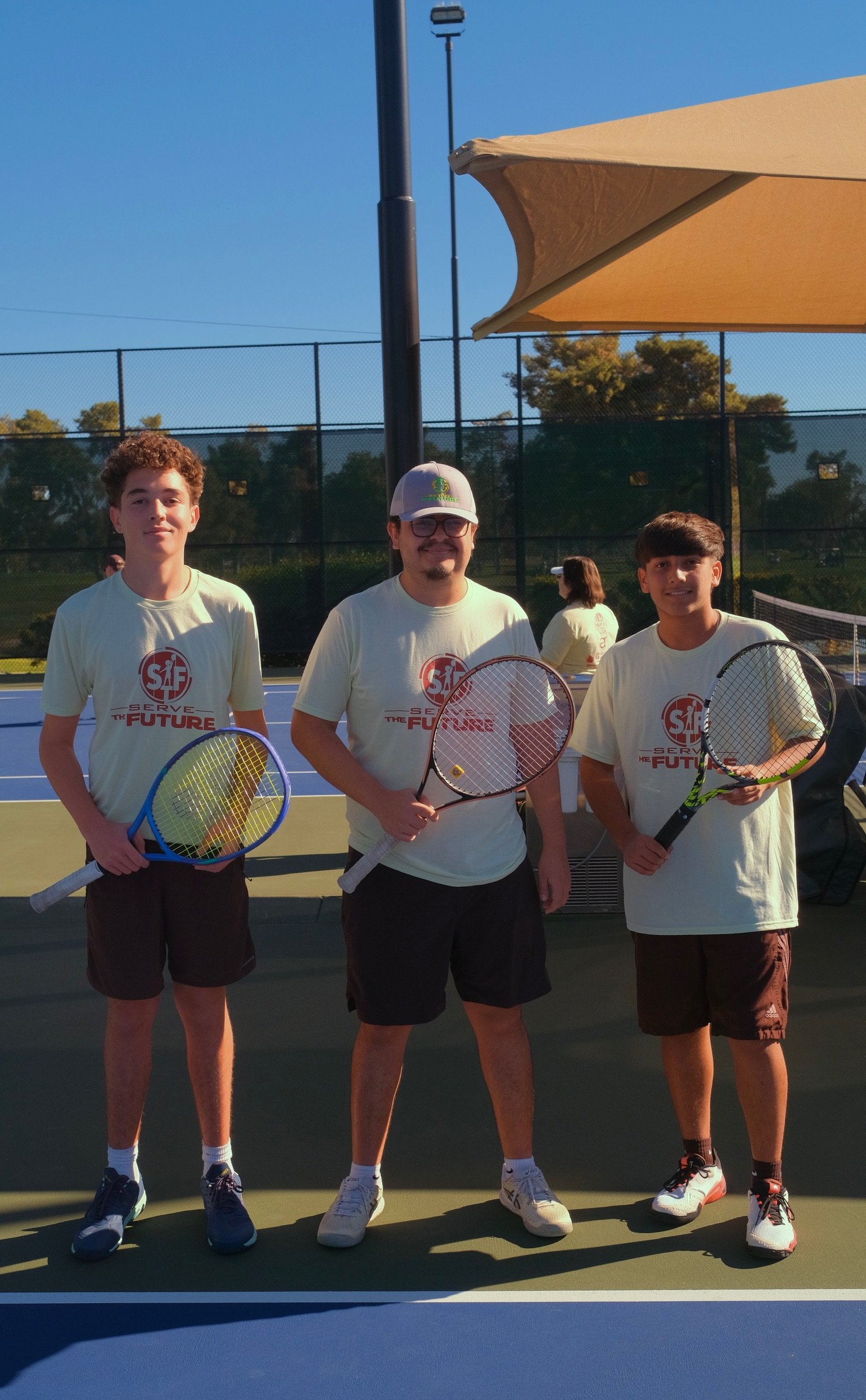 Three young boys standing on a tennis court holding tennis rackets, wearing matching T-shirts with the slogan 'Serve the Future.'