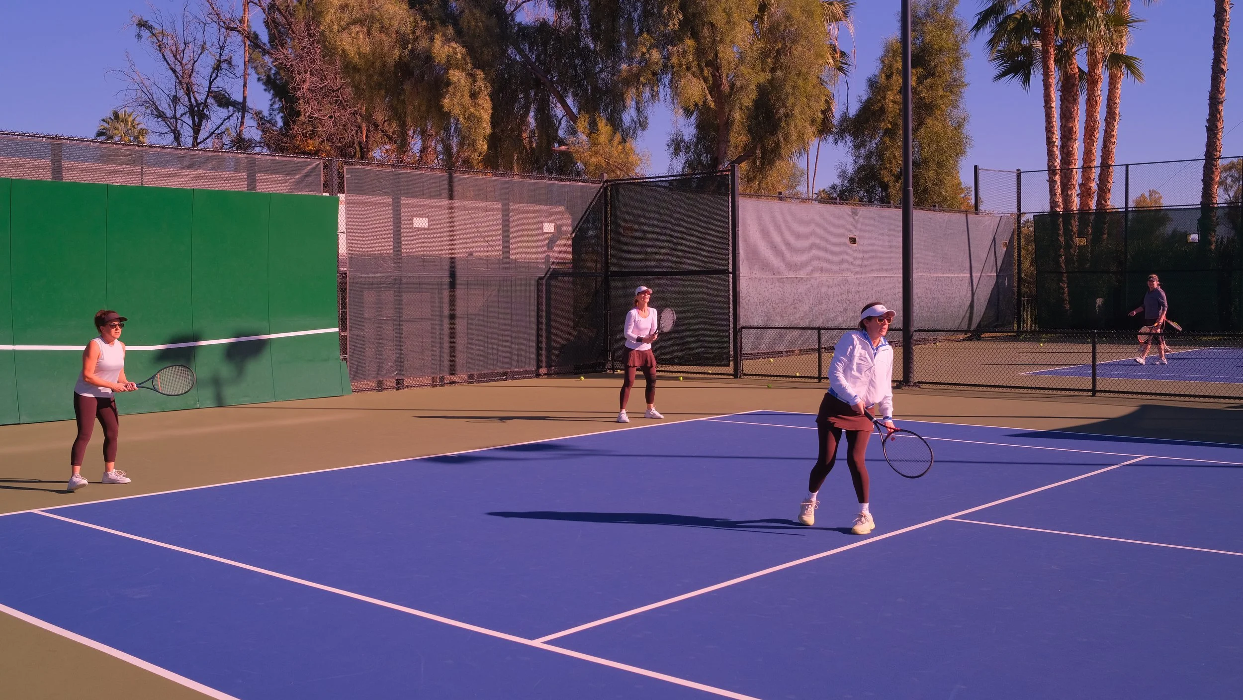 Four women playing doubles tennis on an outdoor court, with two near the net and two toward the back, surrounded by trees and fencing.