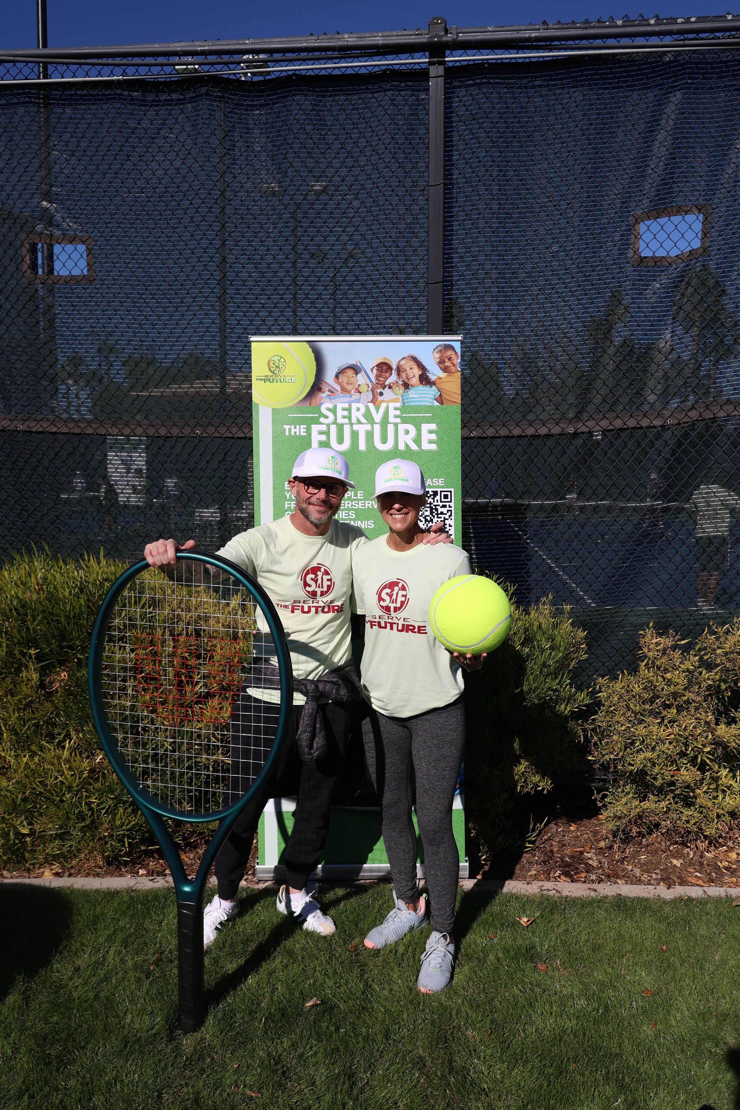 Two people wearing matching t-shirts and caps standing on grass in front of a tennis court, smiling at the camera, with a large tennis racket and a tennis ball.