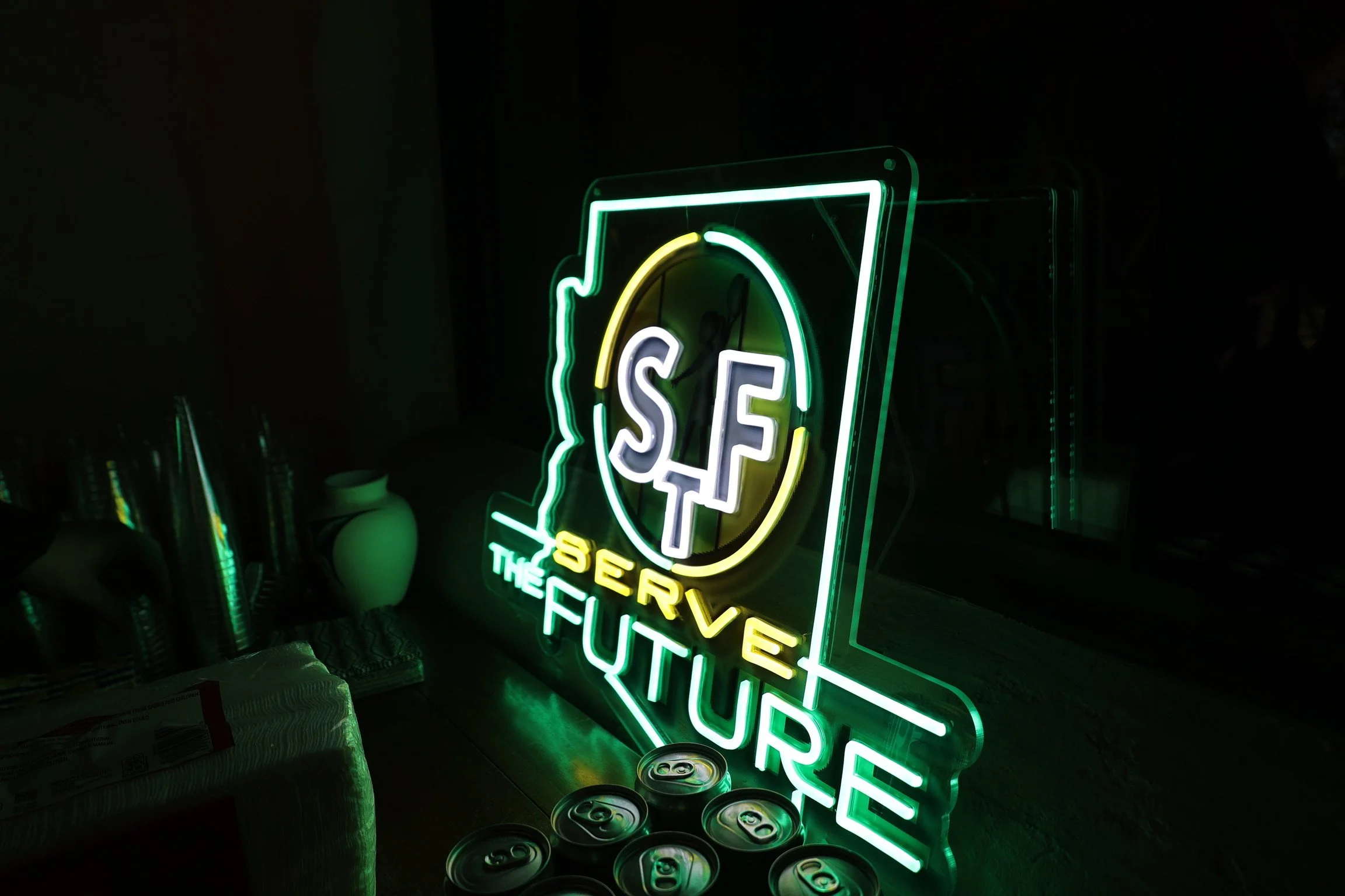 Neon sign reading 'Serve the Future' with a stylized outline of the state of Wisconsin and the letters 'SF' inside a circle, in a dark room.