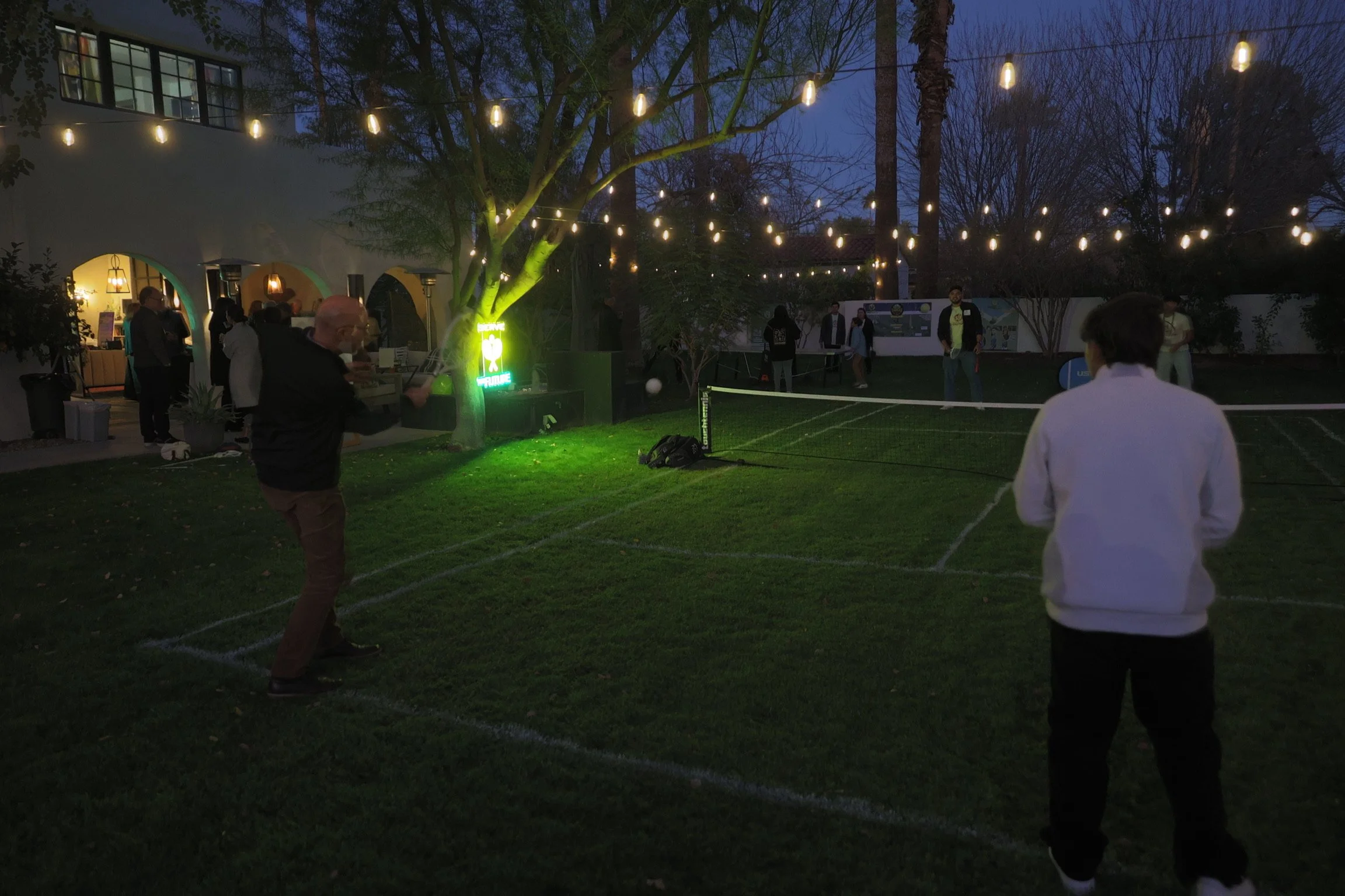 People playing nighttime pickleball on an outdoor court with string lights overhead and a building with open arches in the background.