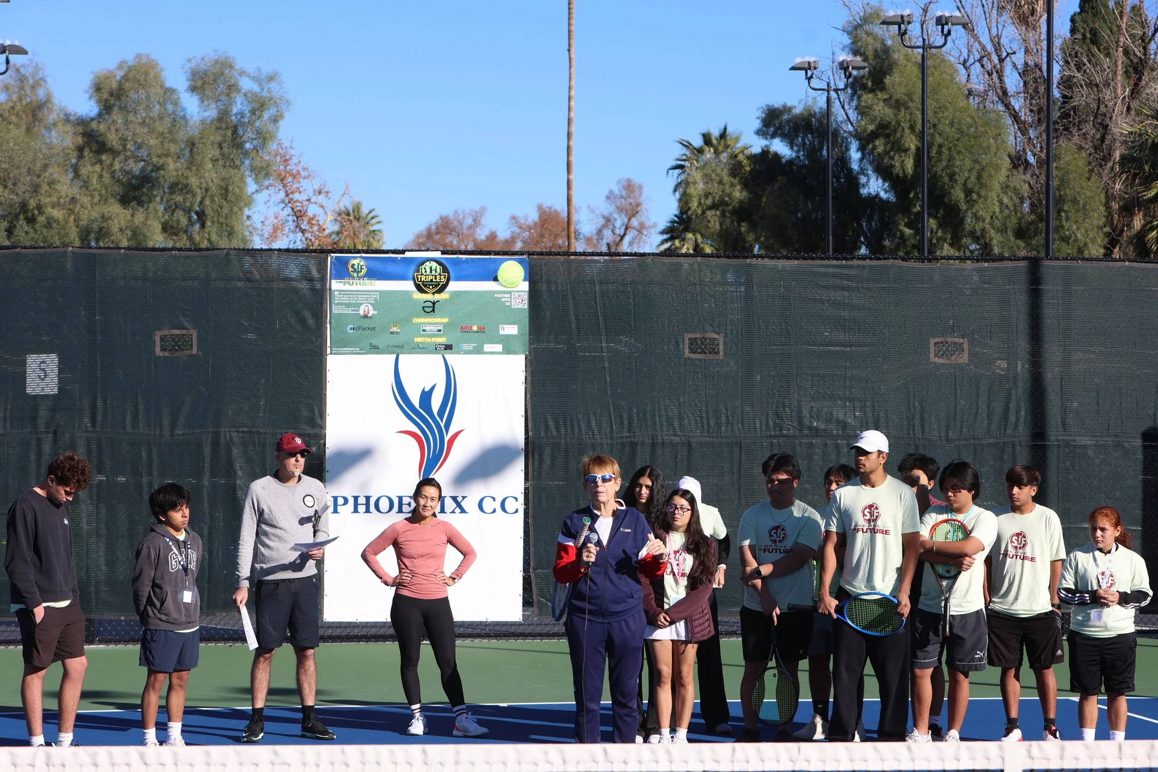 Group of people, including children and adults, standing on a tennis court during an event, with a woman holding a microphone and speaking.