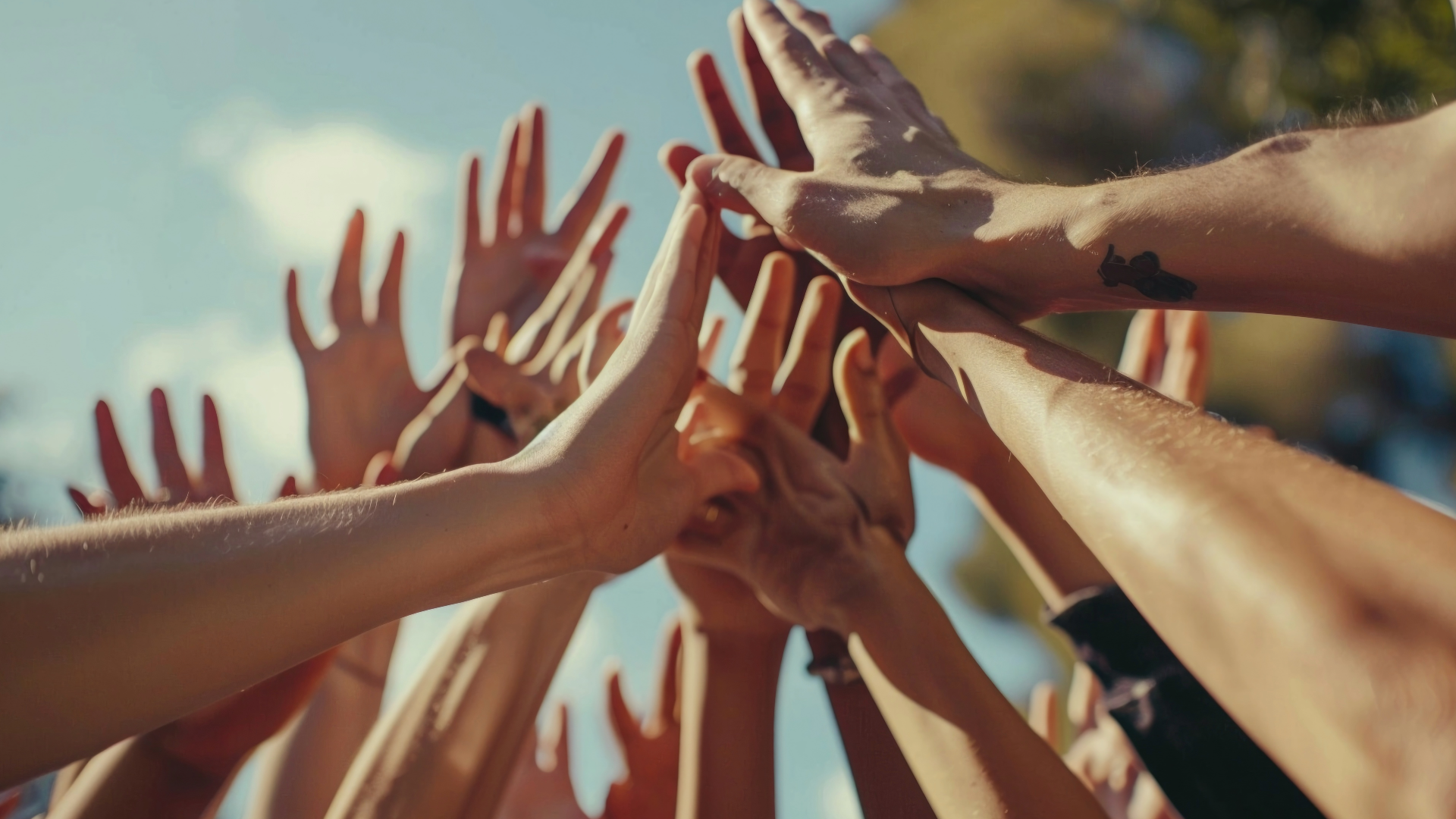 Multiple hands reaching upward and coming together in a high-five gesture with a bright sky in the background.