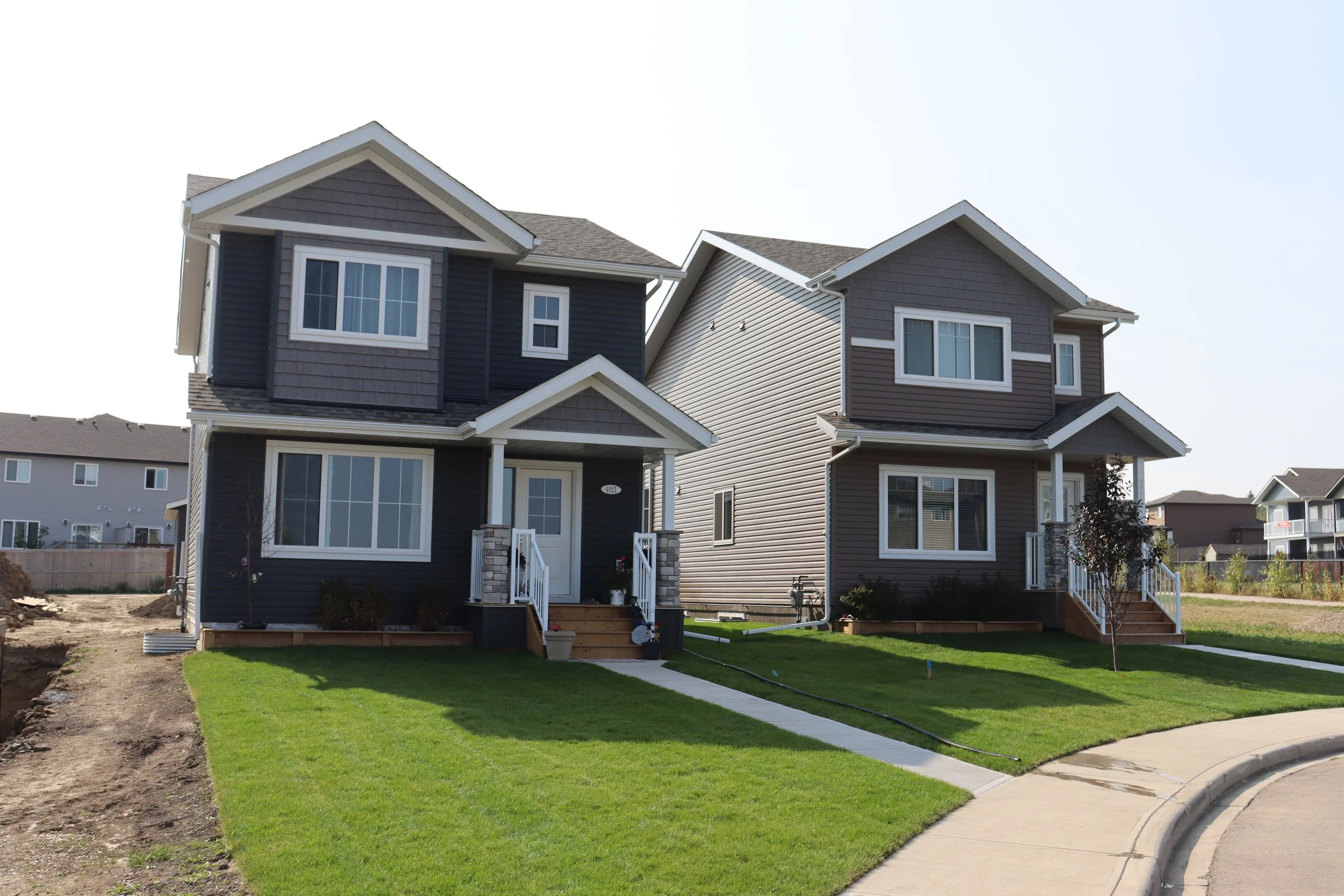 Two new detached suburban houses with manicured lawns and sidewalks, under a clear sky.