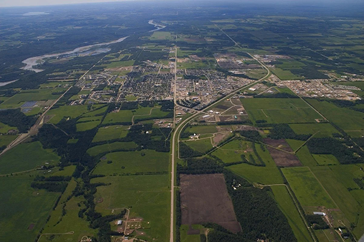 Aerial view of a rural area with fields, forests, roads, and a small town in the distance, with a river flowing through the landscape.