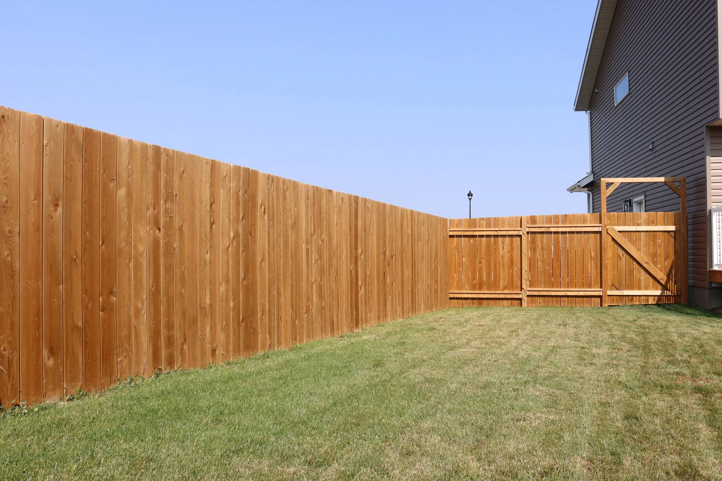 A backyard with a green lawn, a tall wooden privacy fence, and a partial view of a house with siding. A gate in the fence is visible, and the sky is clear and blue.