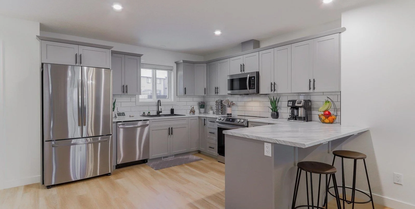 Modern kitchen with stainless steel appliances, gray cabinets, white subway tile backsplash, a window above the sink, and a small breakfast counter with two stools.
