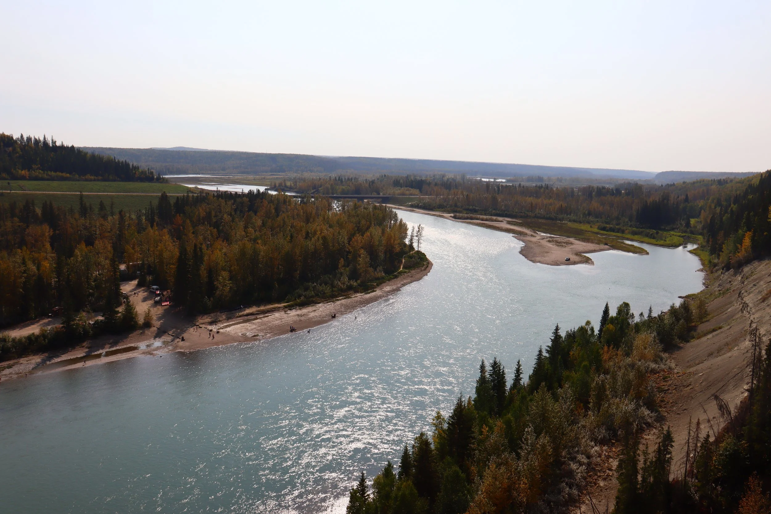 A wide river winding through a forested landscape with trees in fall colors, sandy banks, and a few small boats on the water.