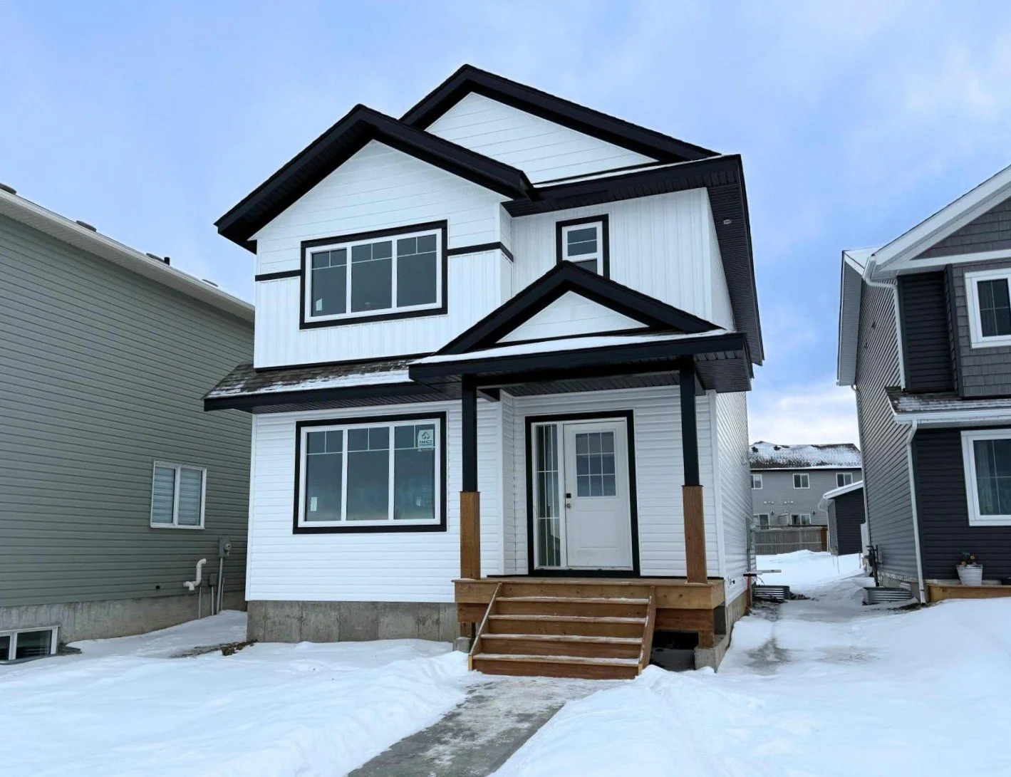 Newly built white and black two-story house with stairs leading to the front door, surrounded by snow, with neighboring houses on either side.