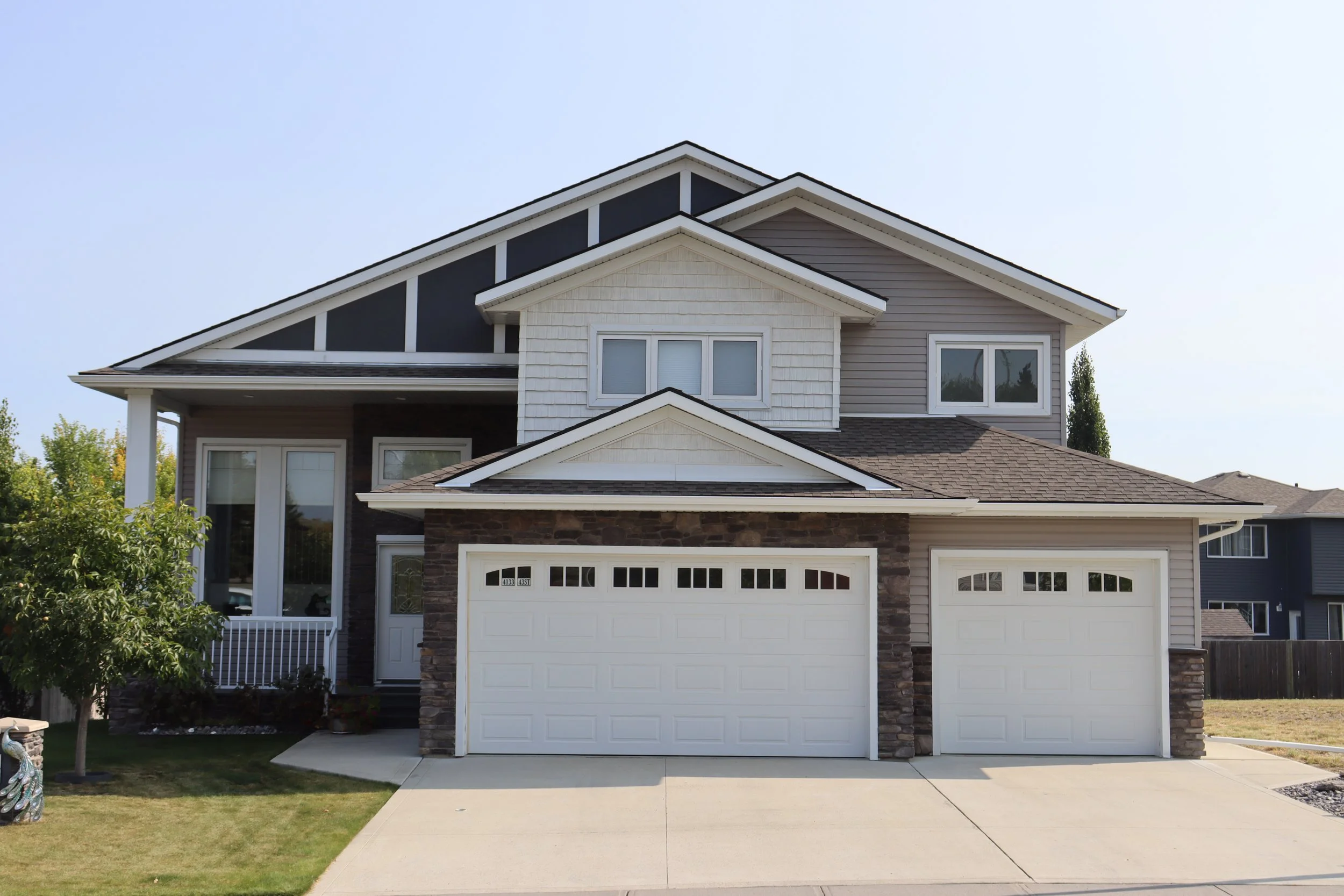 Front view of a two-story modern house with a three-car garage, light brown siding, stone accents, and a small front porch with a tree and lawn.