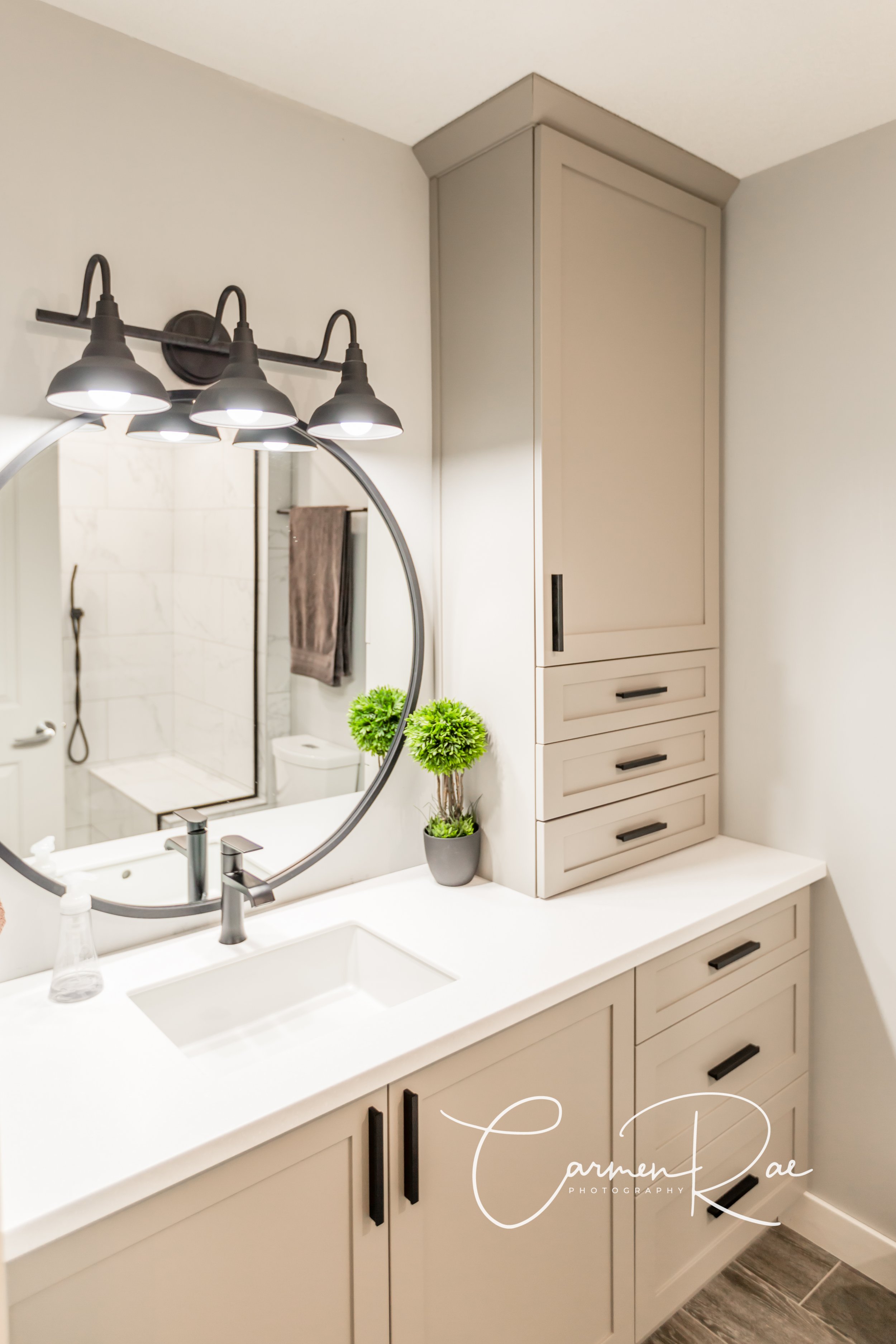 A modern bathroom vanity with a white countertop, black hardware, a vessel sink, a black faucet, a decorative green plant, an oval mirror, and above it, a black three-light fixture. Behind the mirror is a shower with white tiles and a brown towel hanging on a hook.